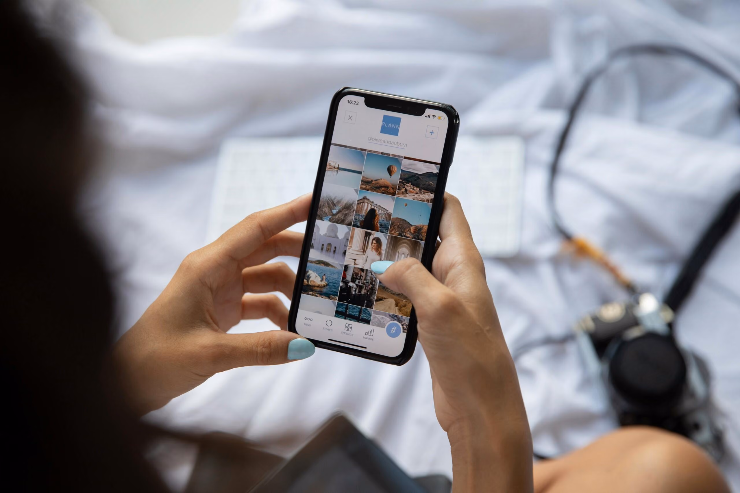 A person holds a smartphone showing a travel photo grid on the PLANN app (@oliveandaburn) with a vintage camera visible nearby.