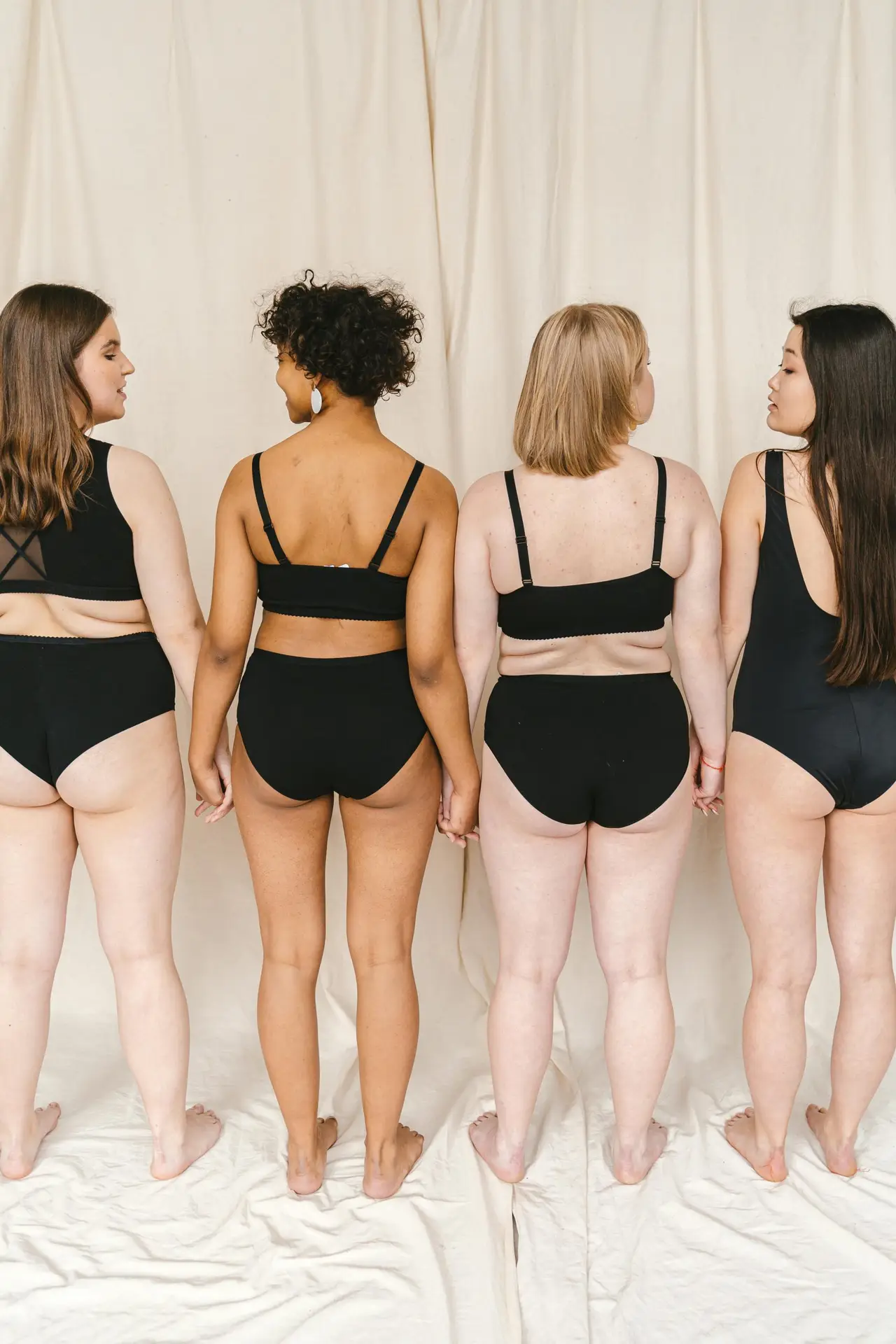 Four women stand back-to-back, holding hands while wearing matching black lingerie against a neutral backdrop.