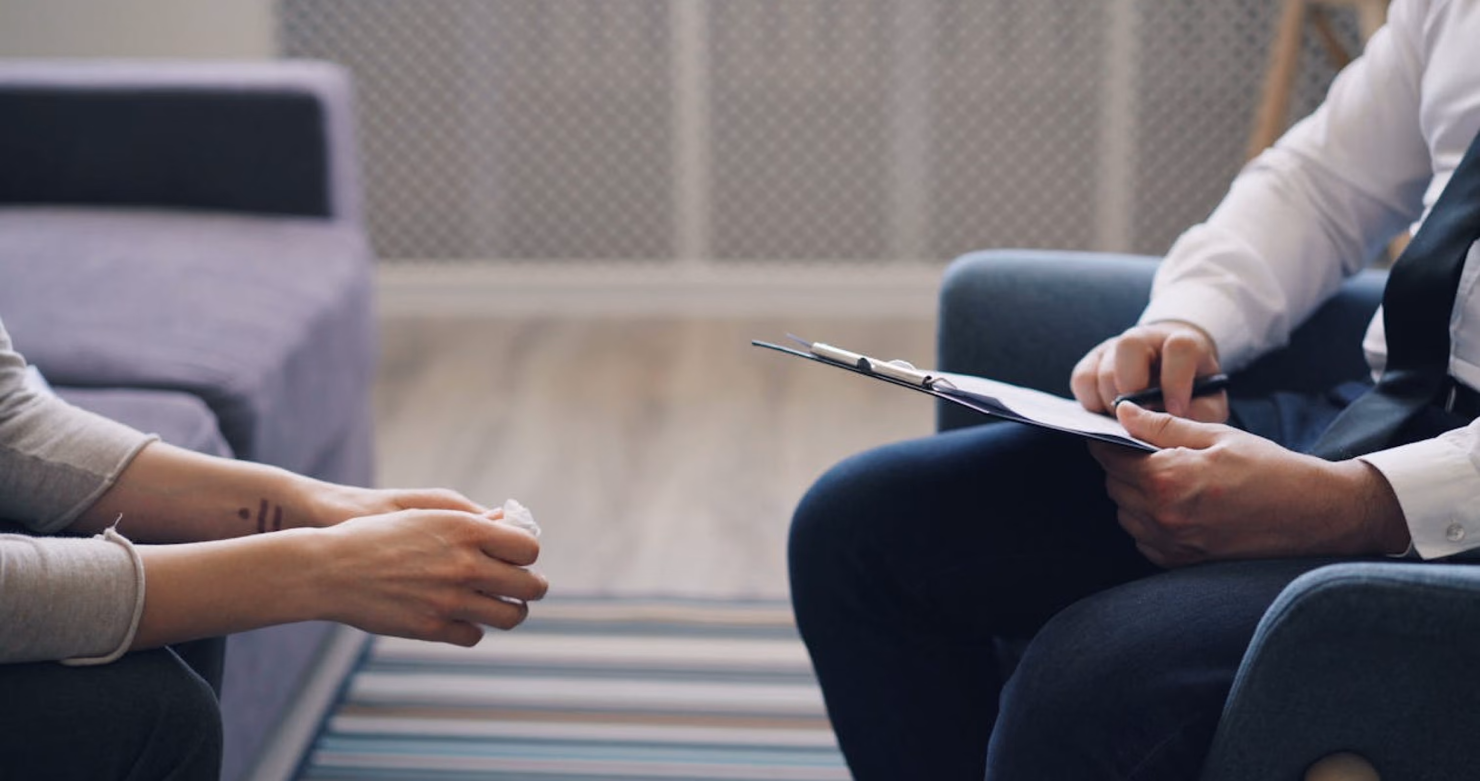 A person writes notes on a clipboard while another holds a tissue during a counseling session.