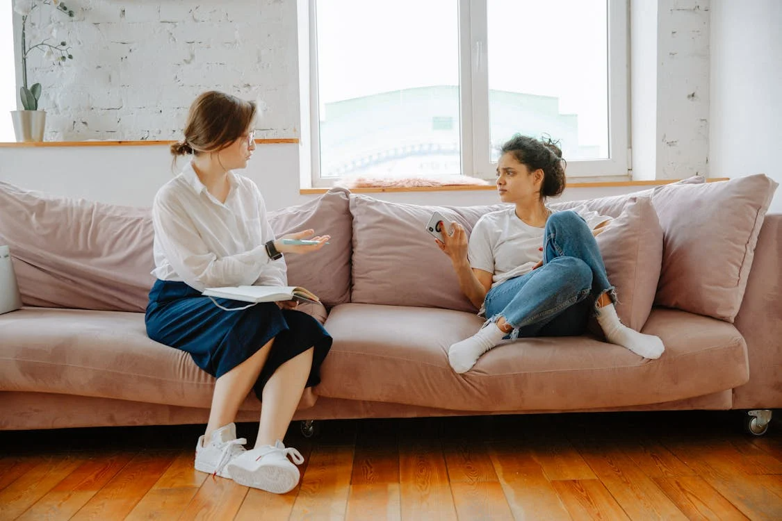 A woman sits on a pink couch holding an open notebook while conversing with another who holds a smartphone displaying the number 6.