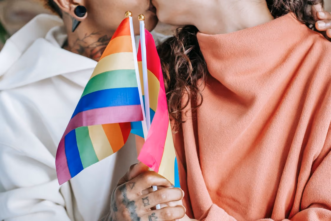Two people kiss while holding small rainbow pride flags.