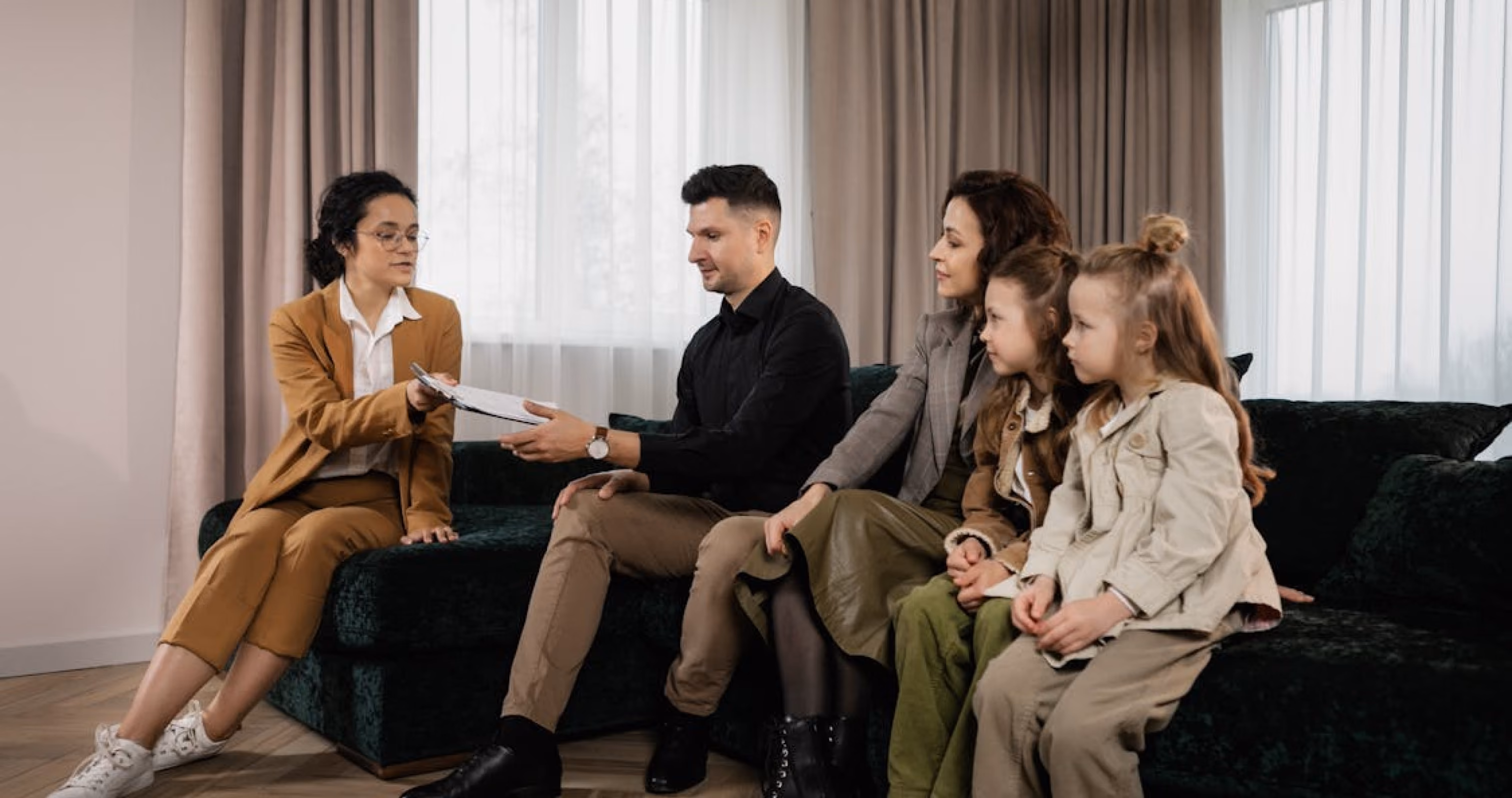 A woman in a tan suit presents documents to a family seated together on a couch.