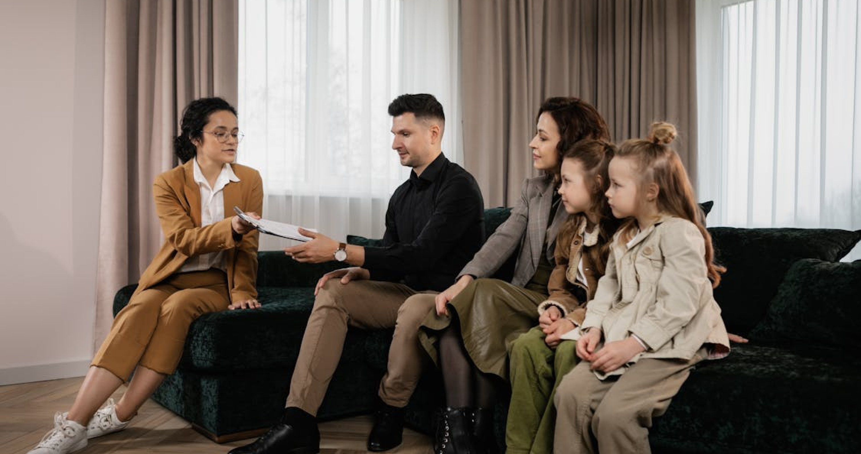 A woman in a tan suit presents documents to a family seated together on a couch.