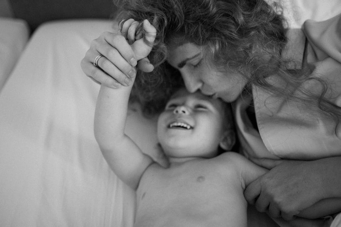 A woman holds a child’s hand and kisses their forehead as they smile.