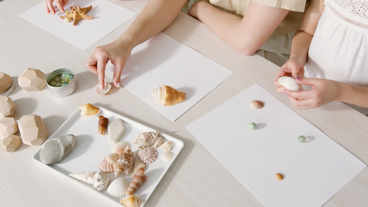 Two people arrange seashells and a starfish on white sheets of paper at a table.