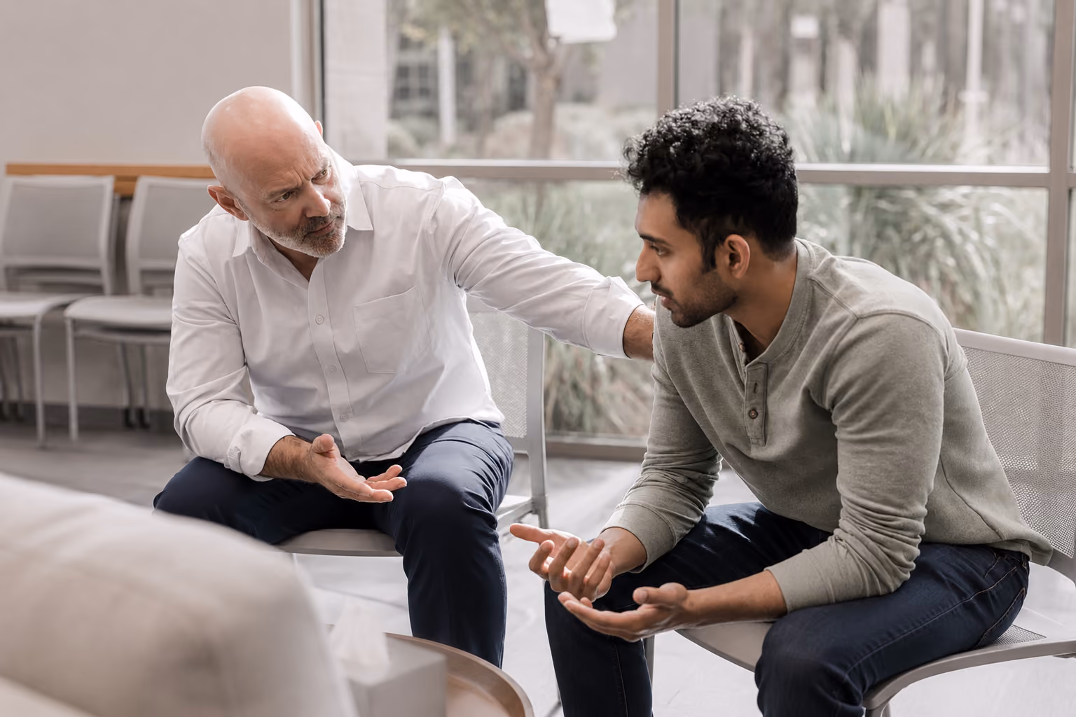 A bald man reaches toward a curly-haired man as they converse with hand gestures while seated indoors.