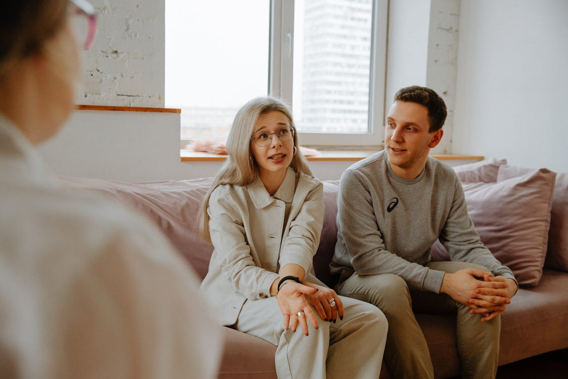 A couple sits on a pink couch, conversing with a therapist.