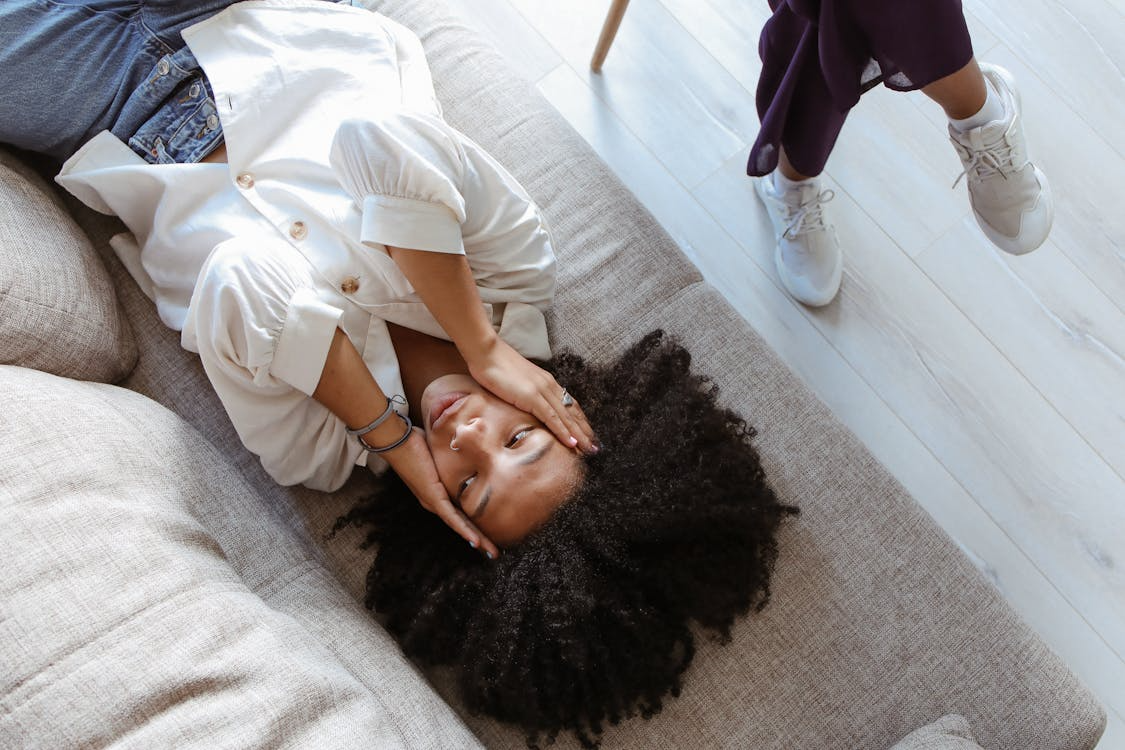 A person lies on a beige couch with hands near their face while another stands nearby wearing white sneakers and purple pants.