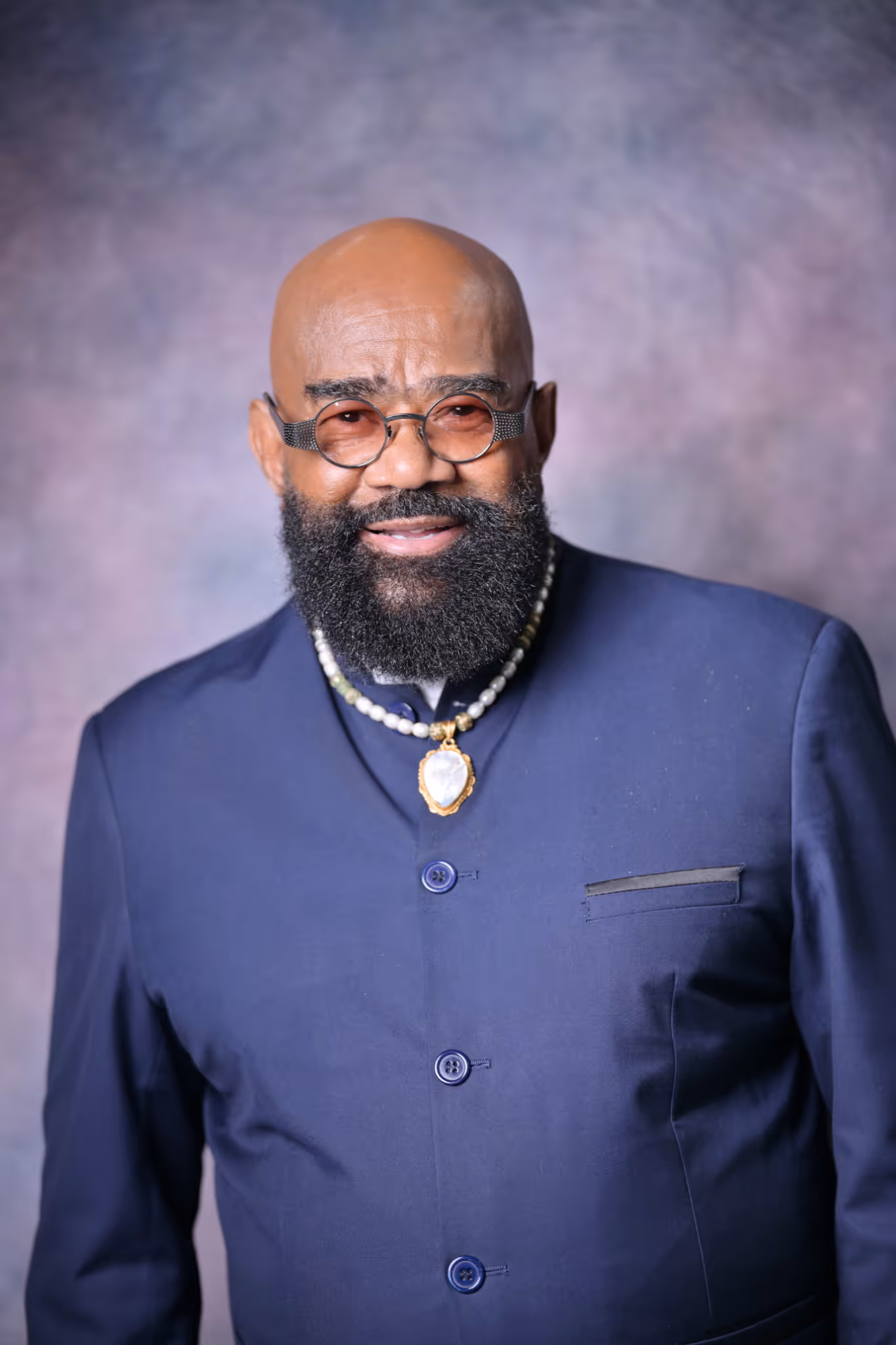 James E. Green wearing tinted glasses, navy jacket, and a beaded necklace with a pendant.