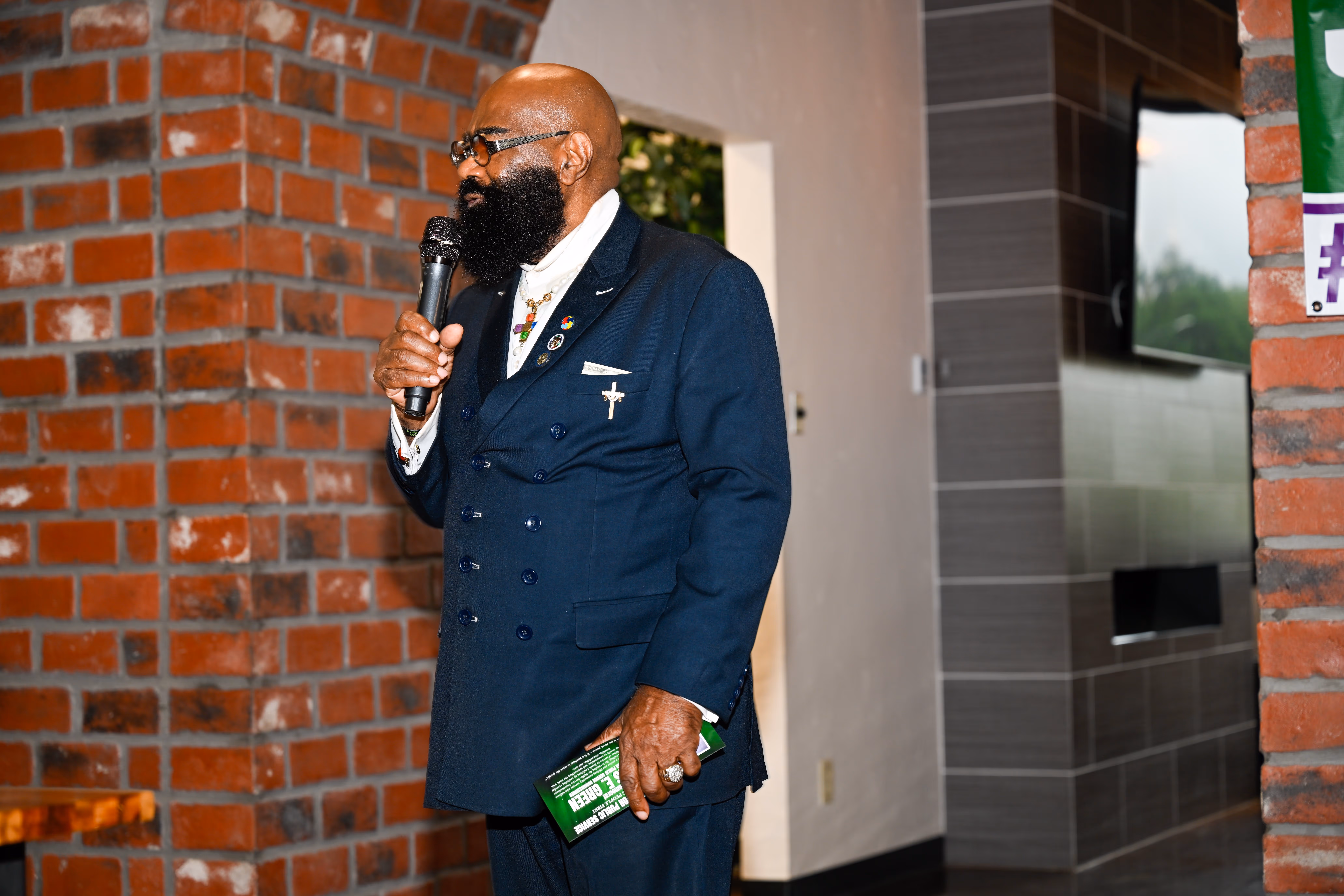 Man in a dark suit speaks into a microphone holding a green booklet by a brick wall.
