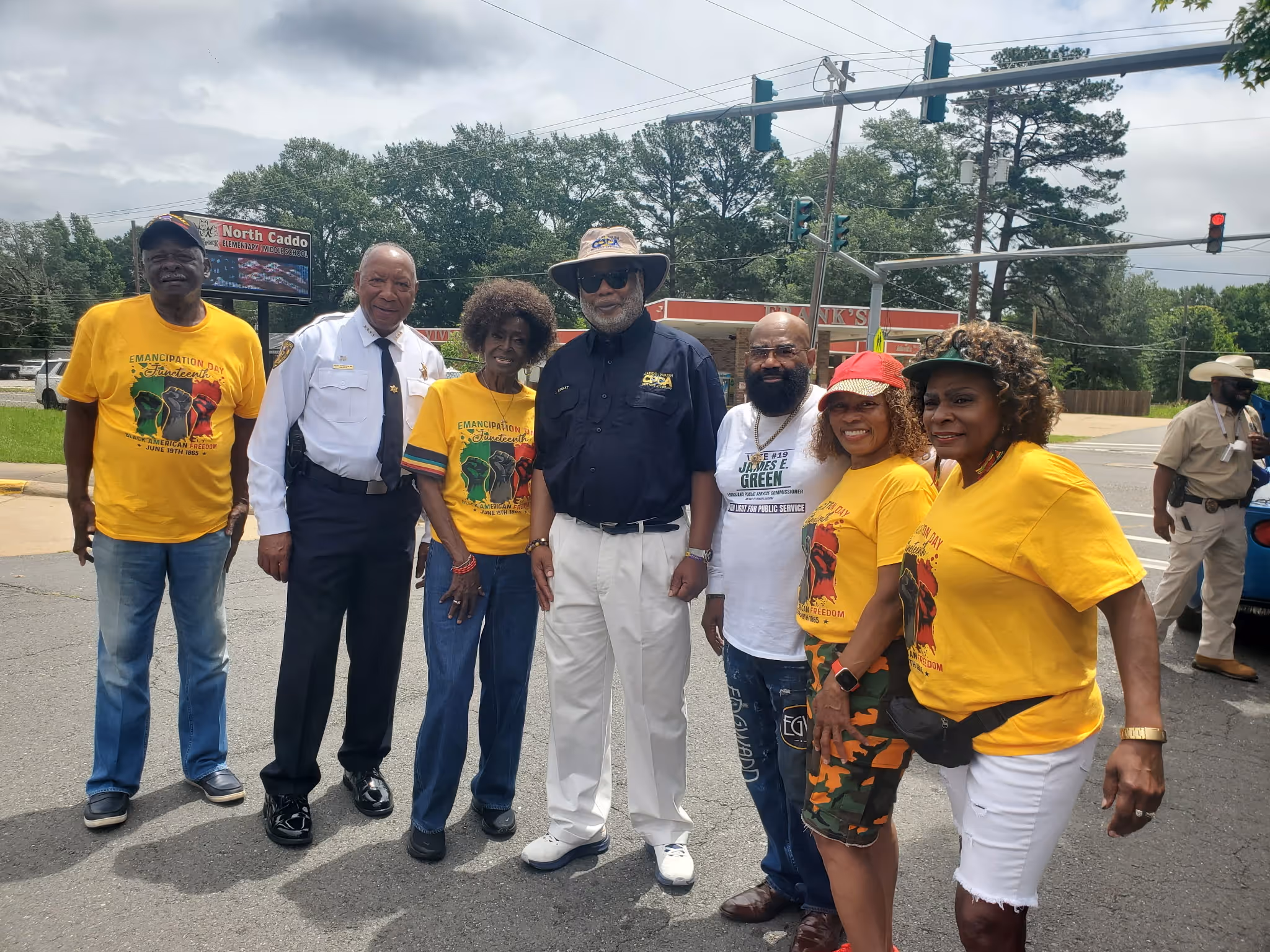 Group of seven adults standing outdoors, some wearing yellow Emancipation Day Juneteenth shirts, one in uniform.