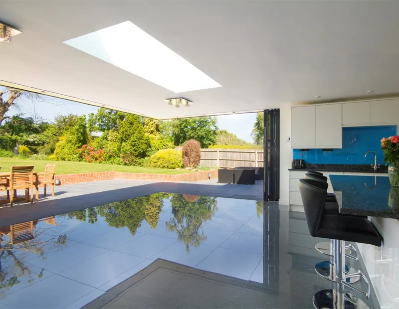 Modern kitchen with black chairs, open wall to garden, and large skylight reflecting on tiled floor.
