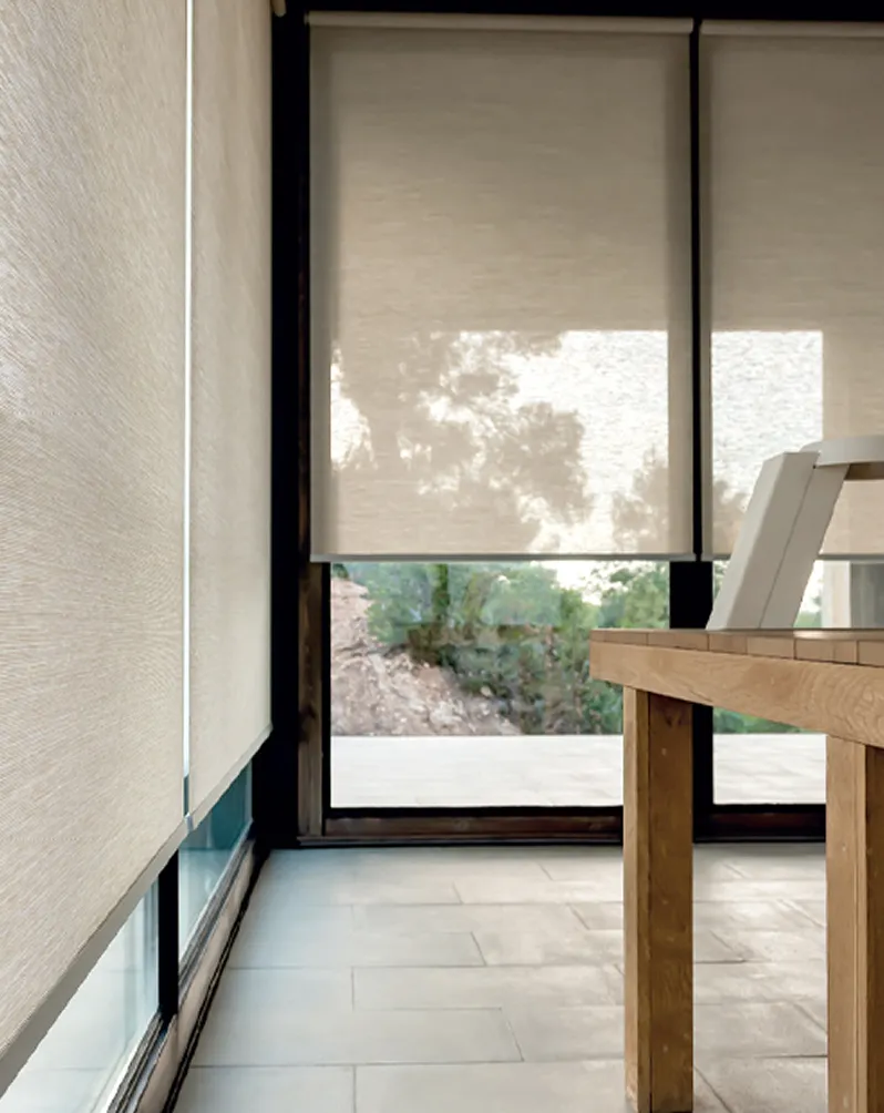 Window with beige roller blinds partially covering a view of trees and a wooden table in front.