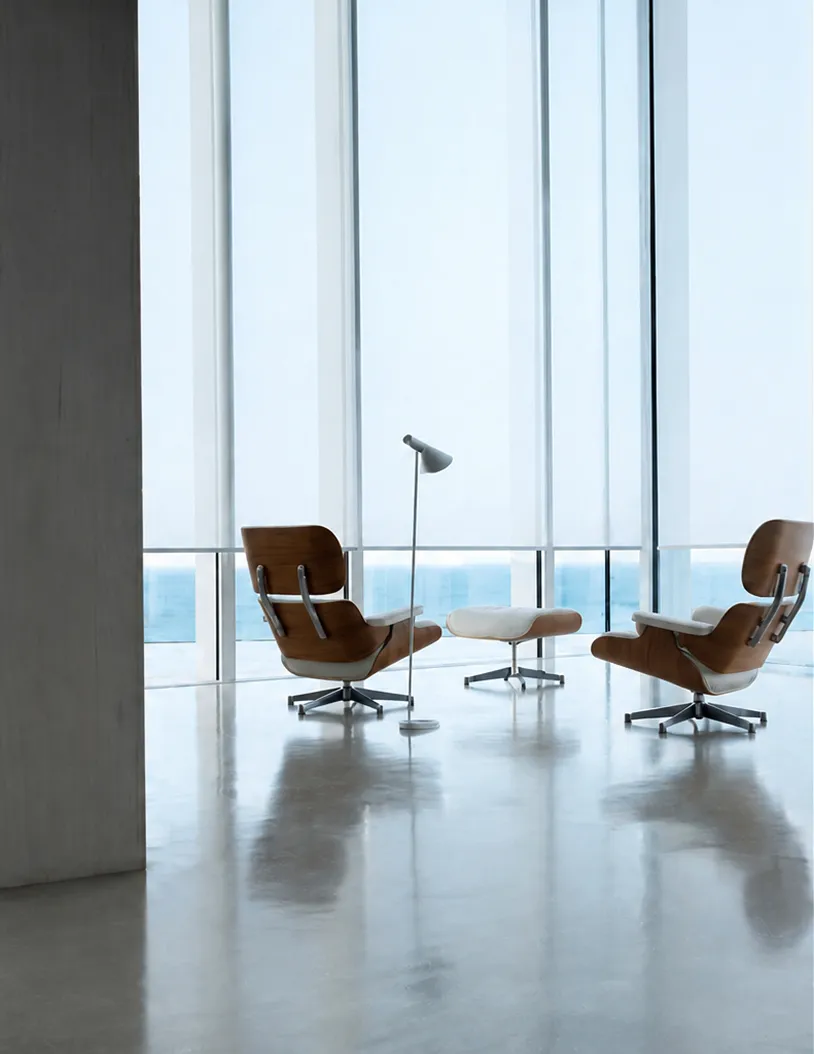 Two modern lounge chairs with ottoman and floor lamp by large glass windows overlooking the ocean.
