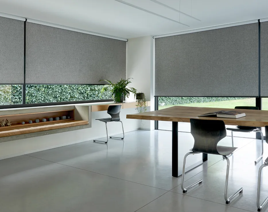 Modern minimalistic room with grey roller blinds, wooden table, black chairs, and green plants by windows.