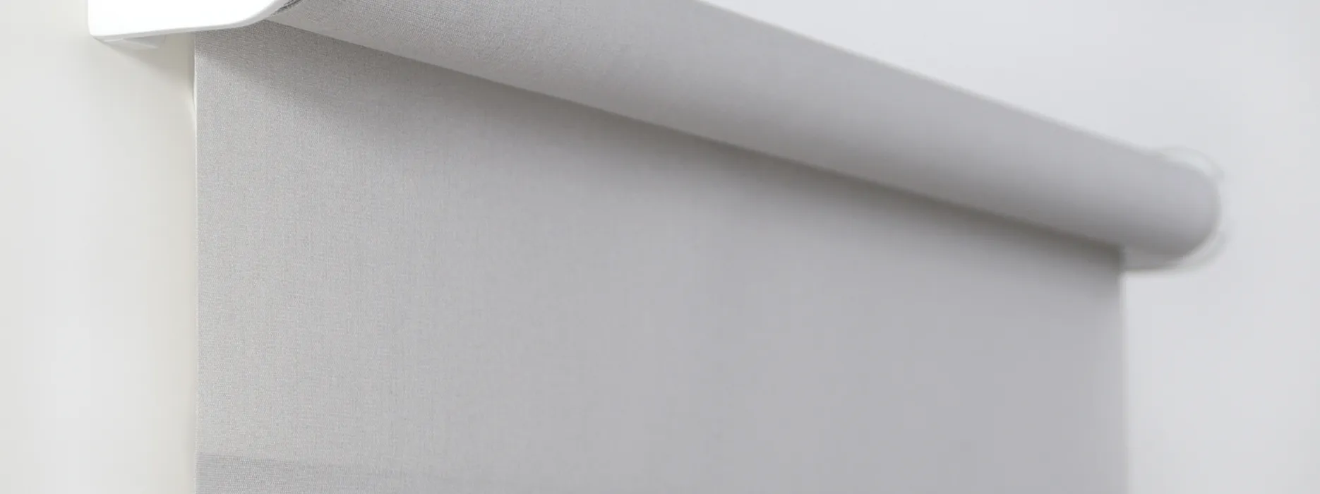 Close-up of a partially rolled down light gray fabric roller blind on a white wall.