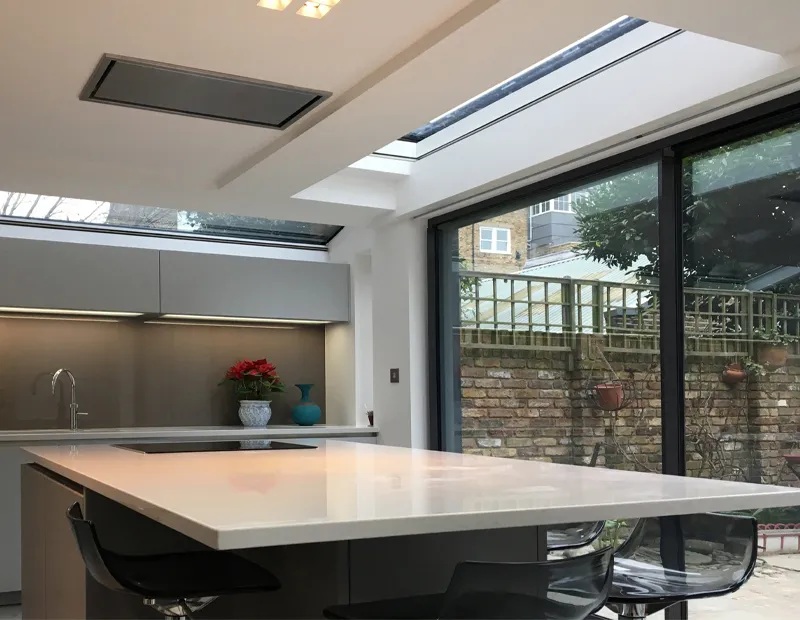 Modern kitchen with large white island, black chairs, skylights, and glass doors showing an outdoor brick wall.