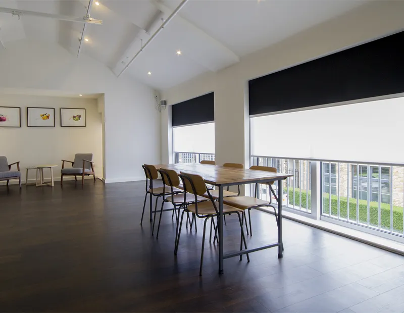 Bright room with a wooden table, six chairs, two armchairs, and large windows with black and white blinds.
