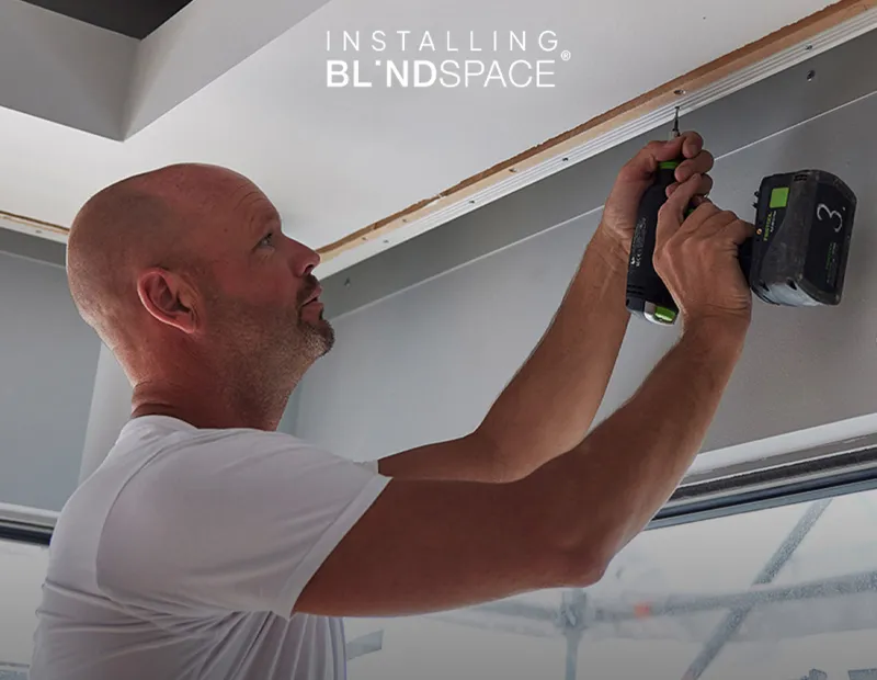 A man in a white shirt installs a Blindspace ceiling panel using a power drill.