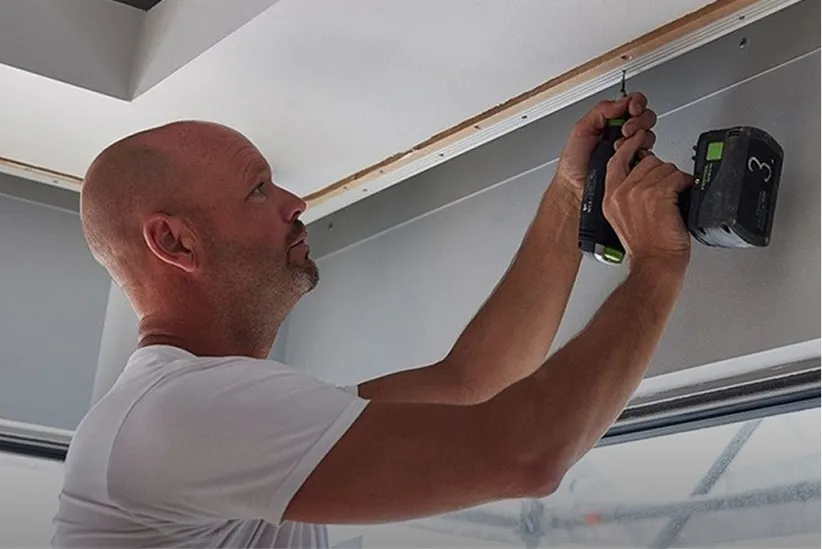 Man using a cordless drill to install a wooden trim near the ceiling indoors.