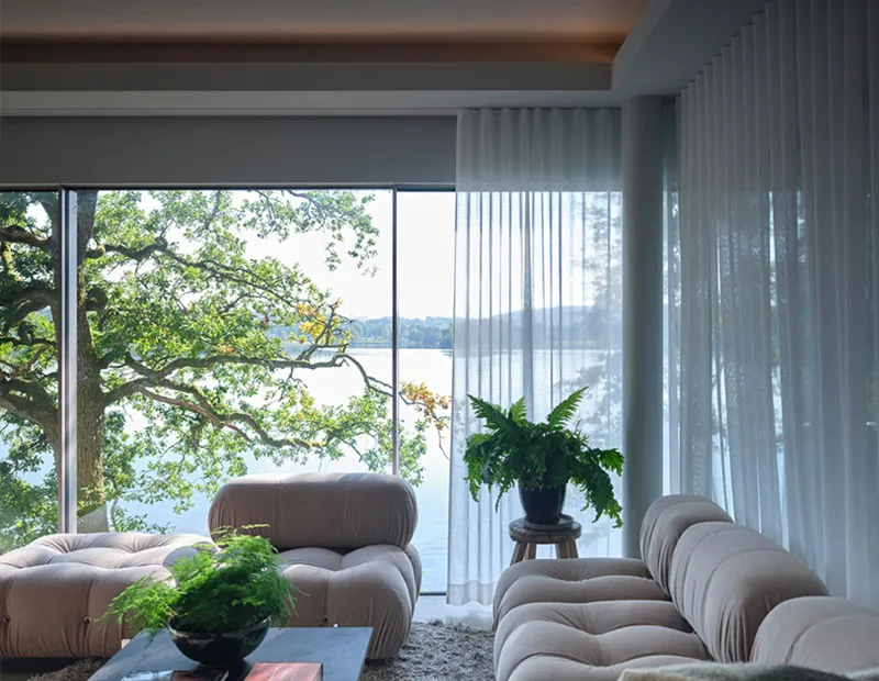 Modern living room with beige tufted sofas, green plants, and large window showing tree and lake view.