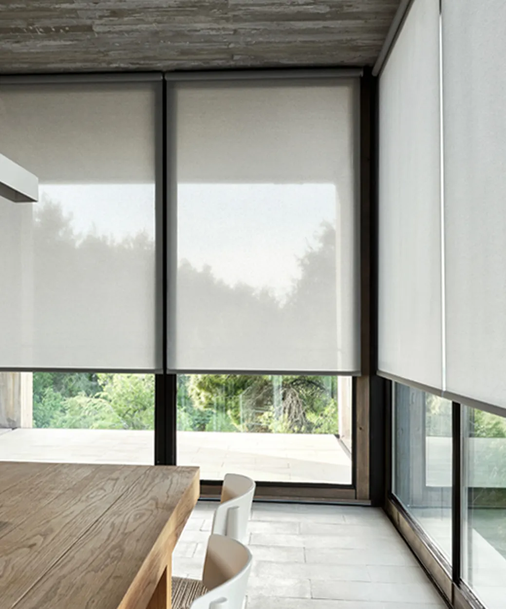 Modern dining area with large windows covered by semi-transparent roller shades and white chairs.
