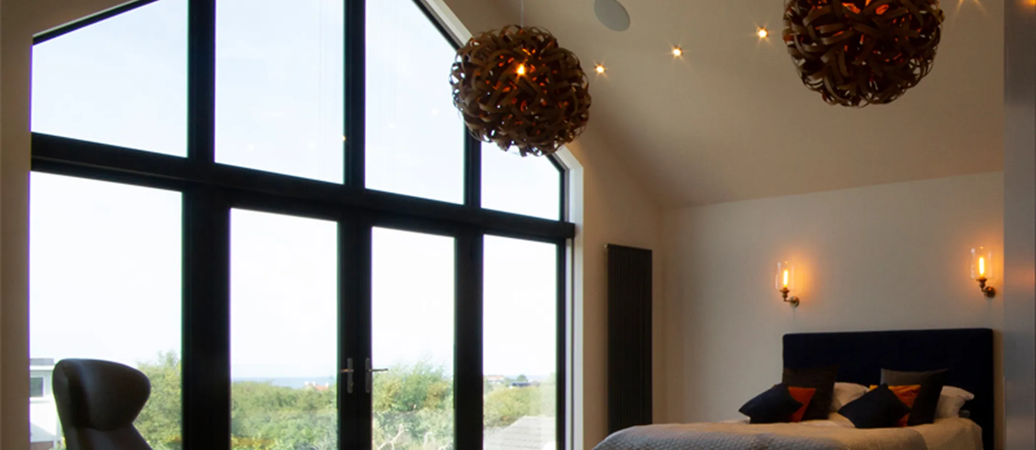Modern bedroom with tall black-framed windows, hanging wooden light fixtures, and a bed with dark pillows.