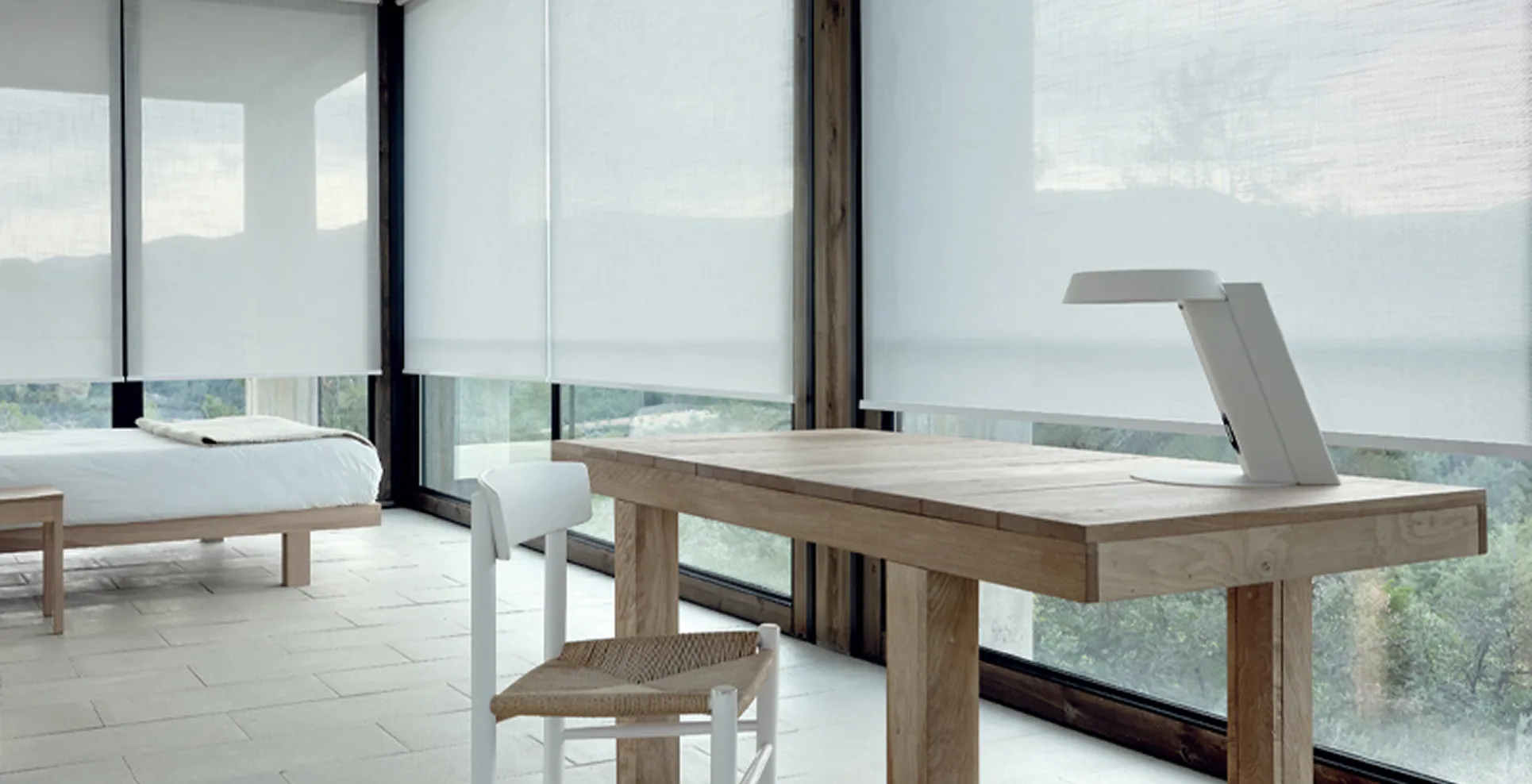 Minimalist room with a wooden table, white chair, bed, and large windows with white roller shades.