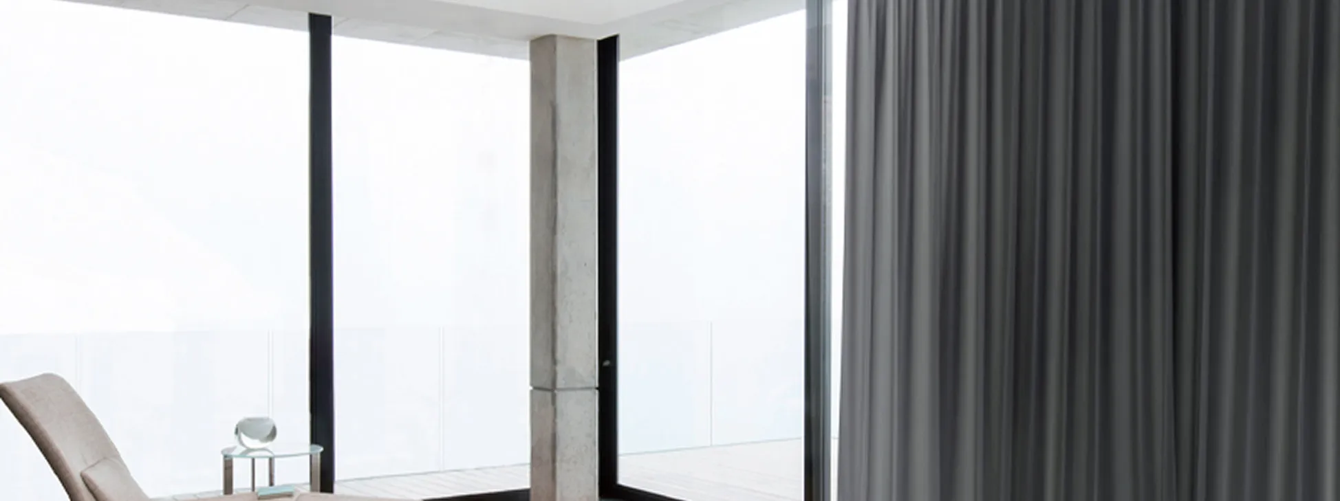 Minimalist room with floor-to-ceiling windows, beige lounge chair, small glass table, and gray curtains.