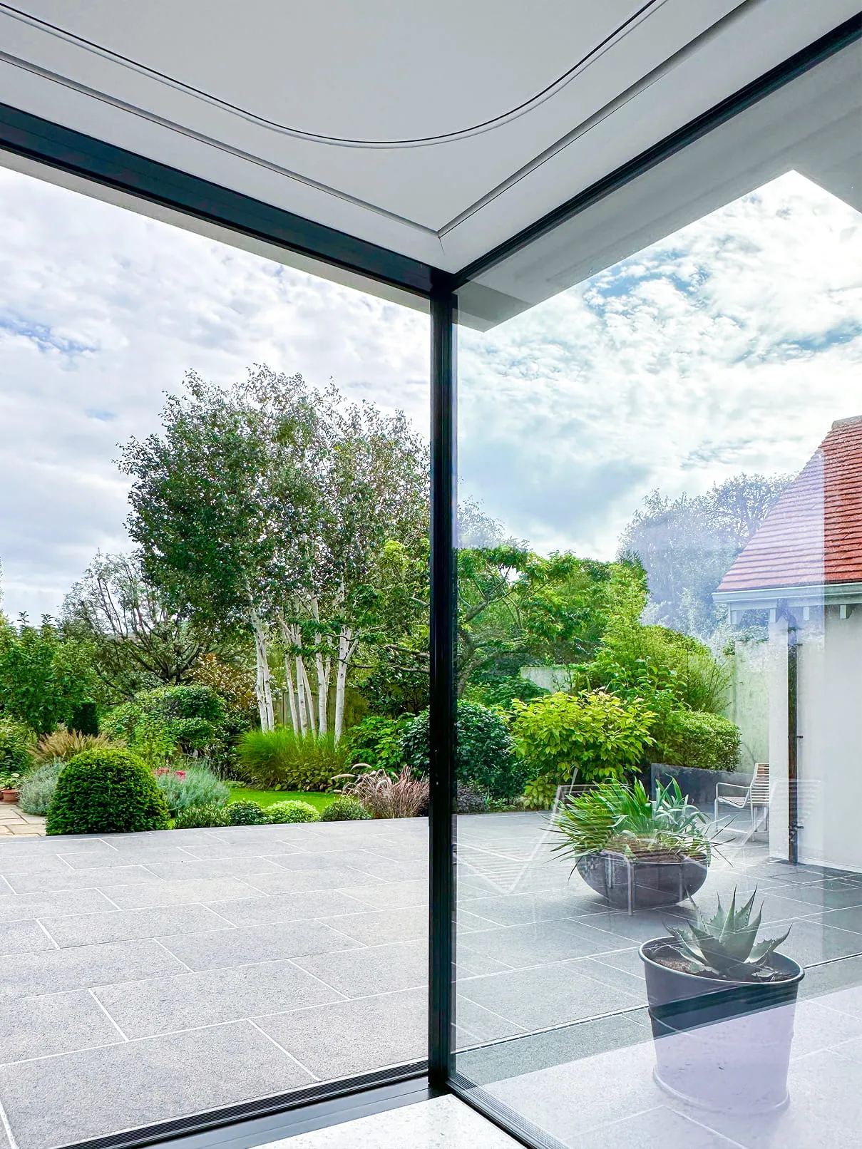 Corner glass walls showing a tiled patio with potted plants and a lush green garden beyond.