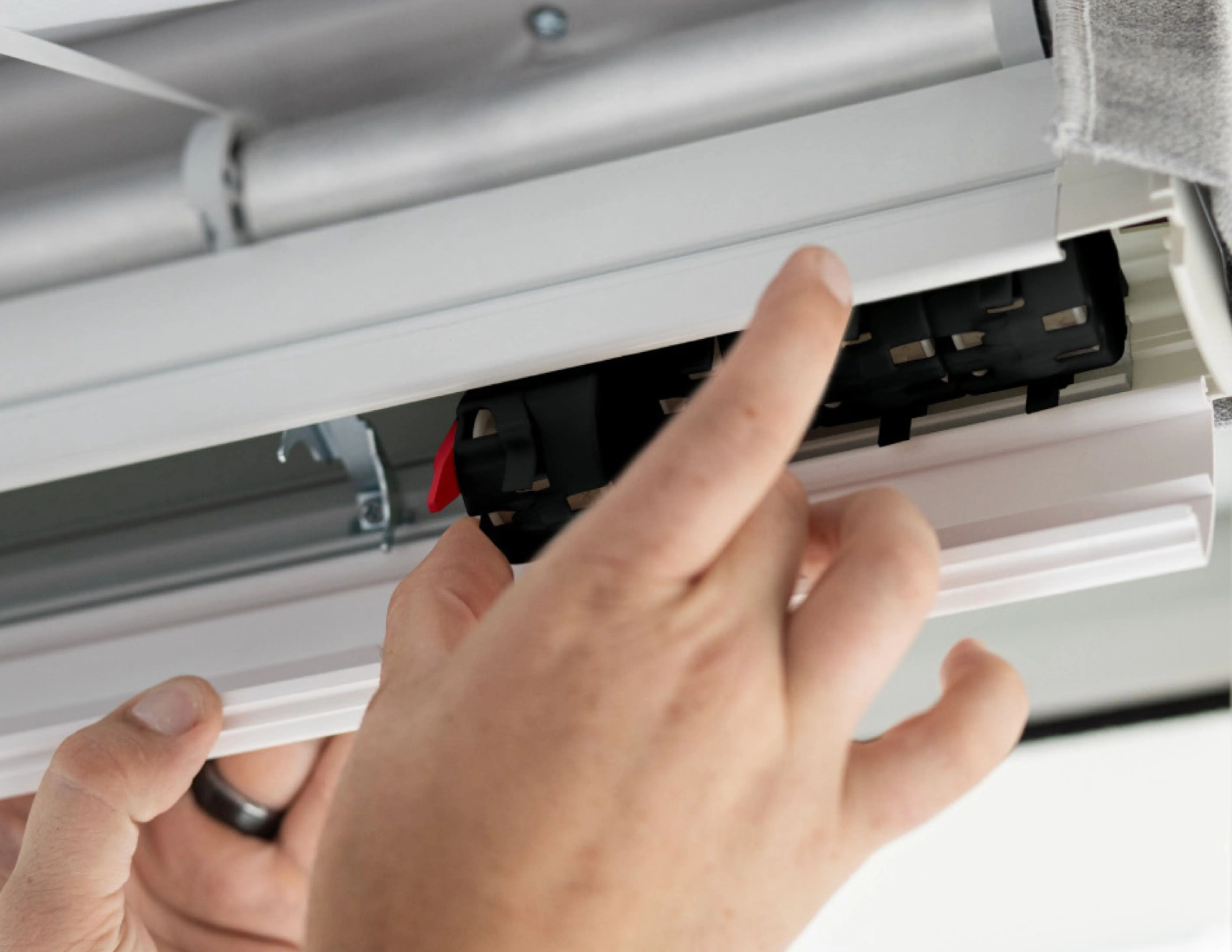 Hands installing or adjusting a white window blind mechanism with a red clip inside.