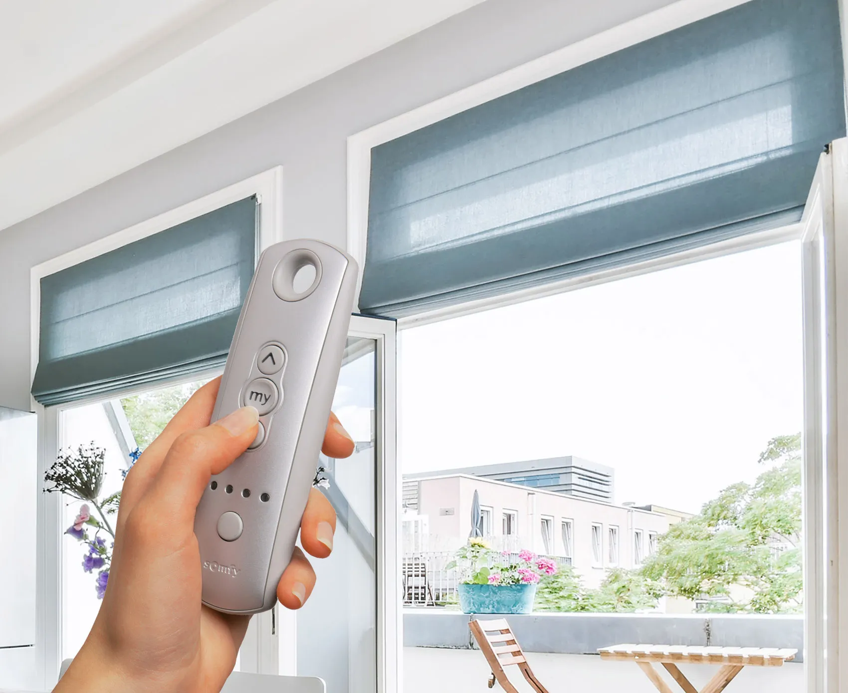 Hand holding a silver remote controlling blue window blinds in a modern home with outdoor view.