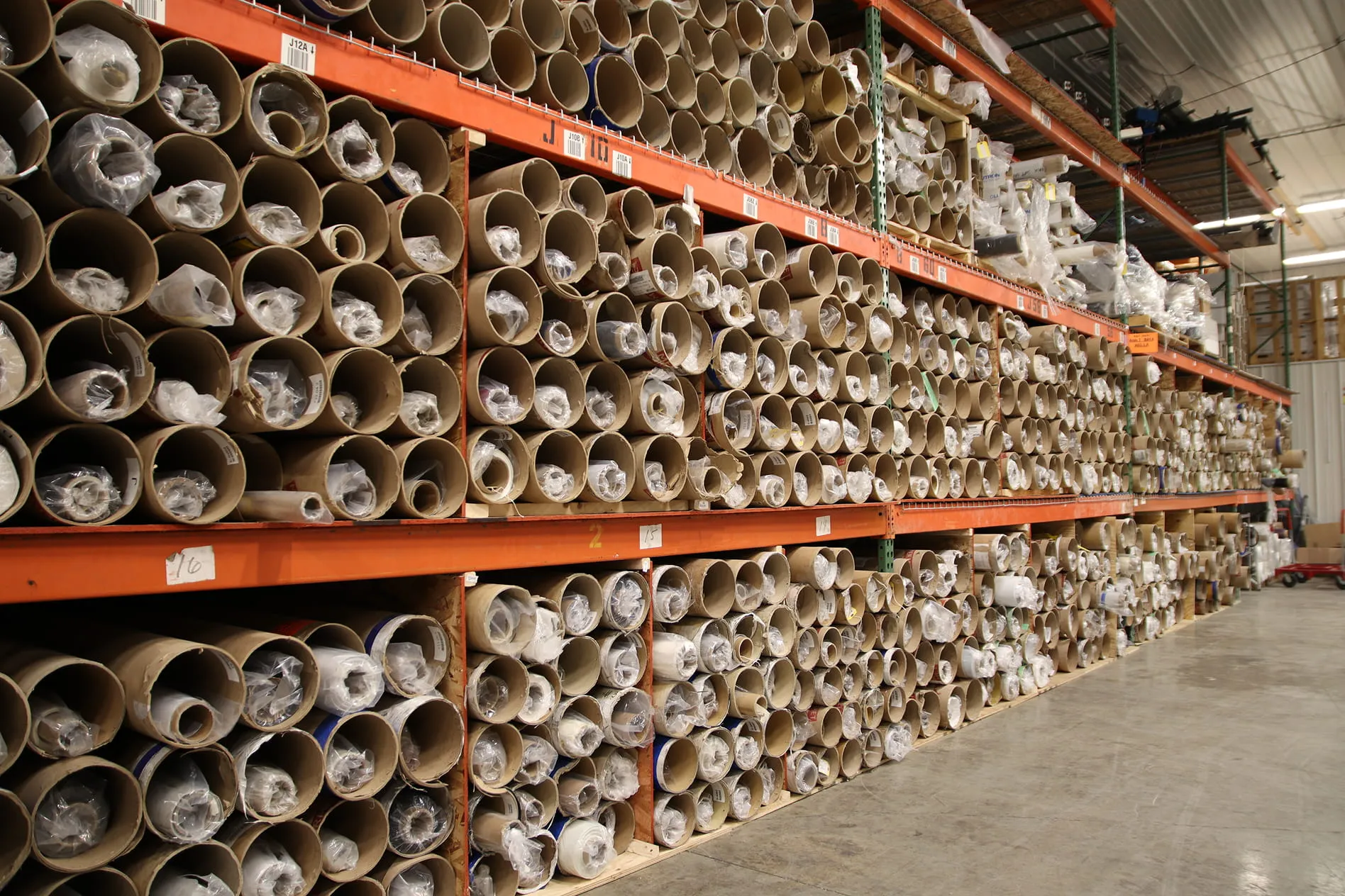 Warehouse shelves filled with large tubes holding rolled materials in plastic wrap.