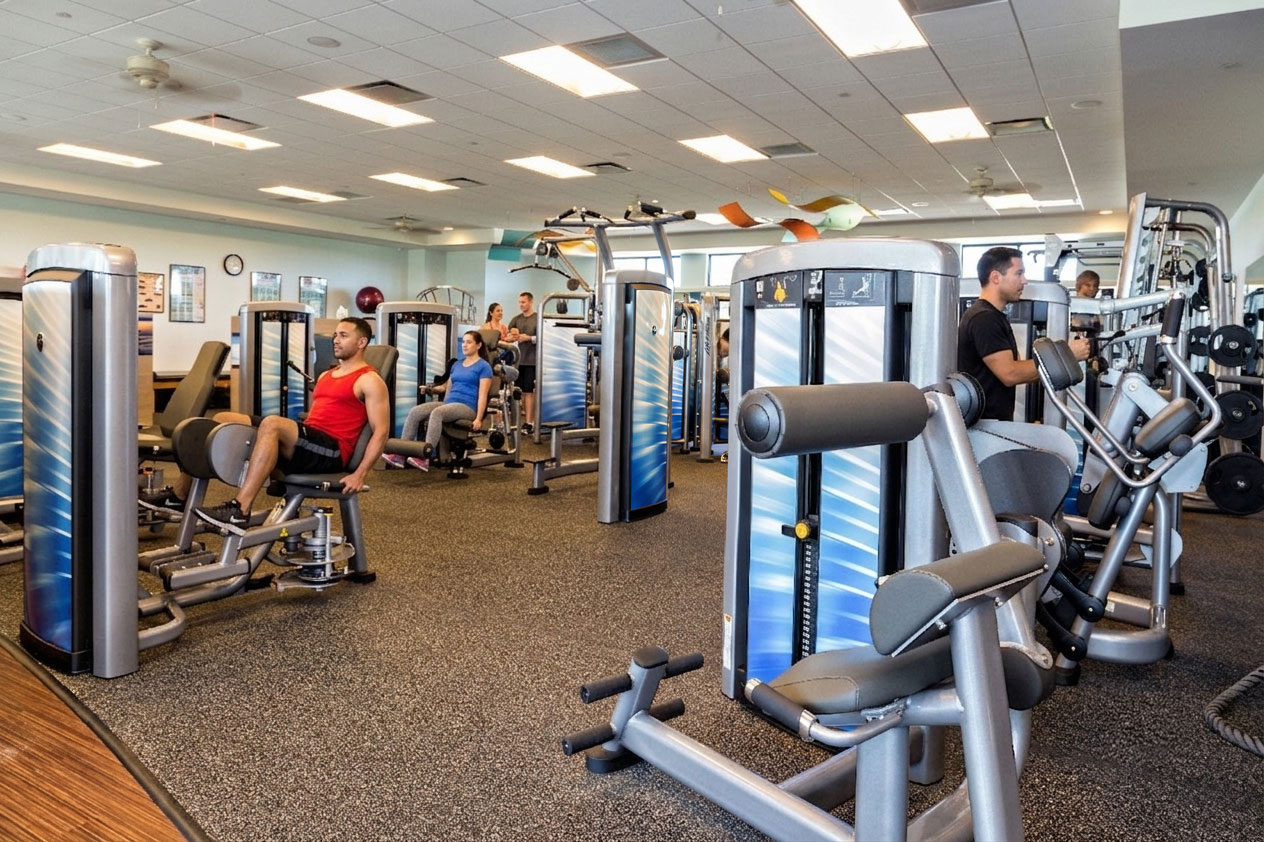 People working out on various strength training machines in a spacious gym with modern equipment and bright overhead lighting.