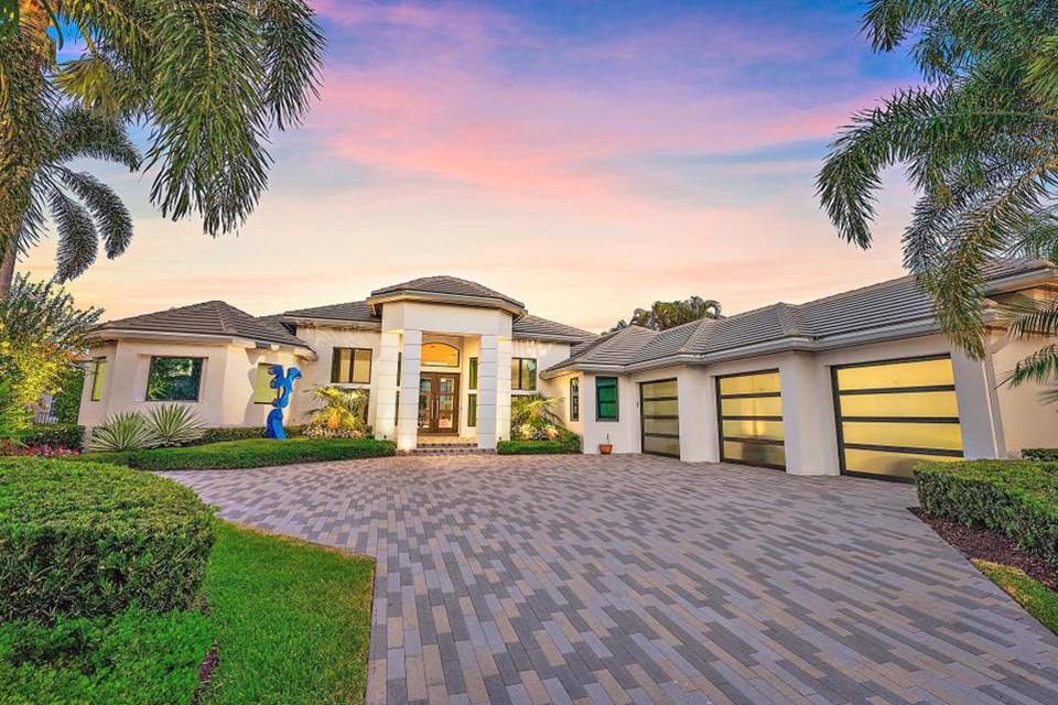 Modern single-story house with three garage doors, a paved driveway, palm trees, green shrubs, and a colorful sunset sky.
