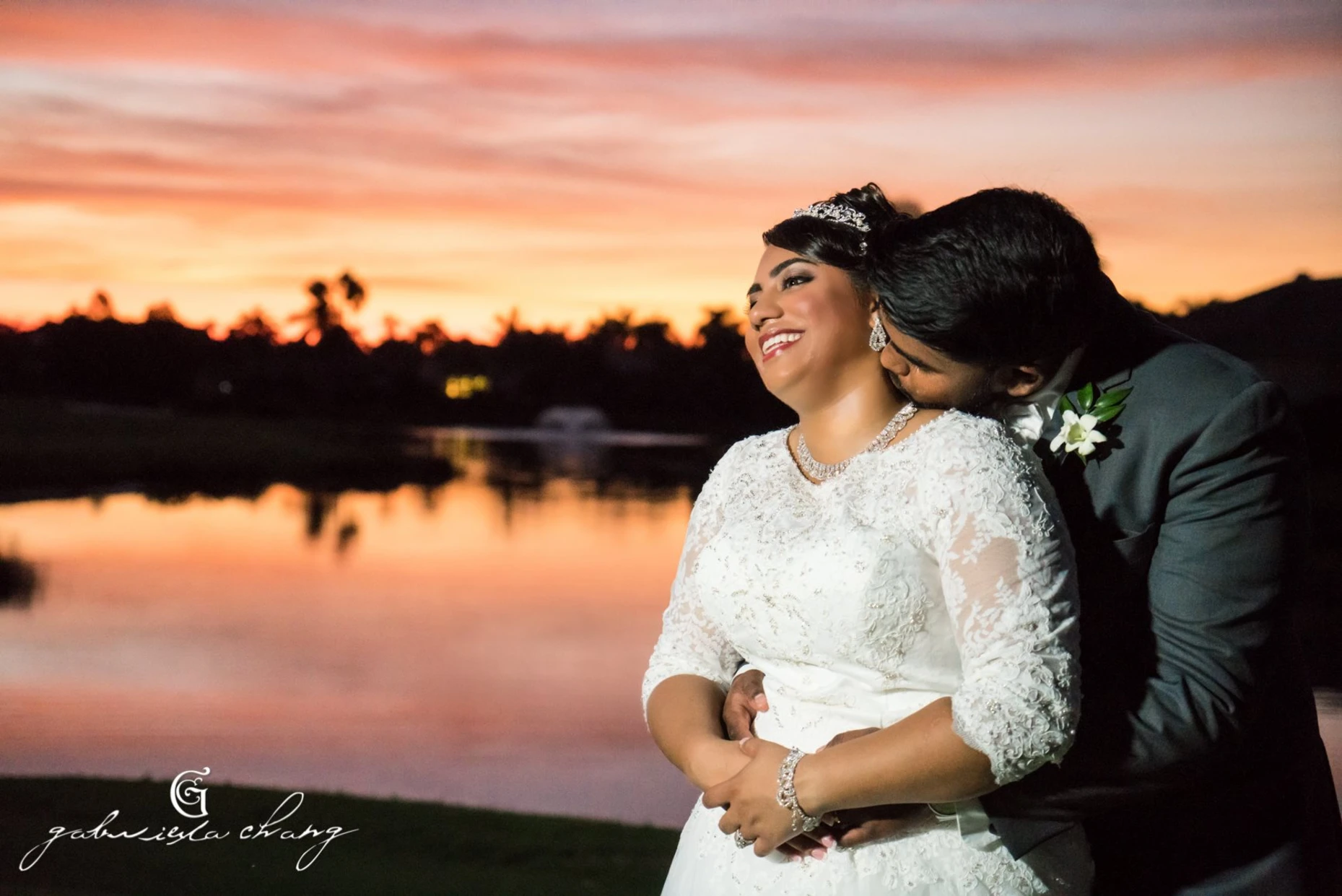 Bride and groom in wedding attire embracing and smiling by a lake at sunset with colorful pink and orange sky.