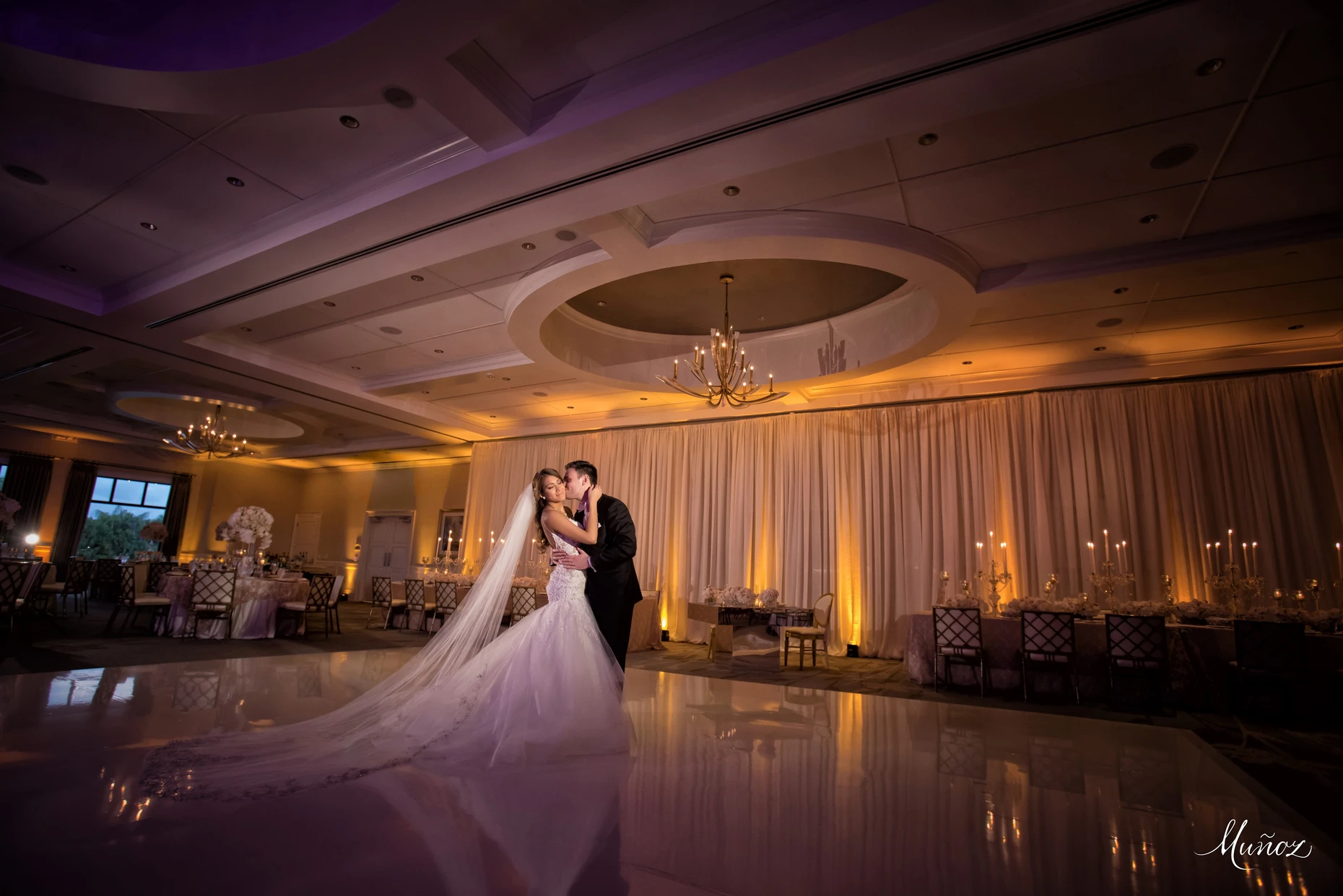 Bride and groom dancing closely on a polished dance floor in a warmly lit, elegant wedding reception hall with draped curtains, chandeliers, and candlelit tables.