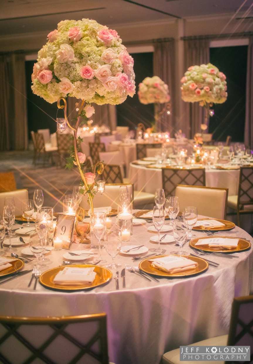Elegant round banquet table set for an event with gold-rimmed plates, folded napkins, multiple wine glasses, and silverware, topped with a tall floral centerpiece of white and pink roses with small hanging candles, surrounded by matching tables with similar decorations in a warmly lit room.