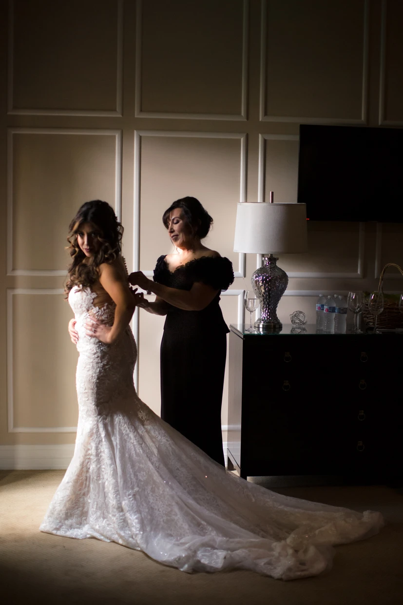 Bride in a fitted lace wedding gown with a long train stands with hands on hips while a woman in a black dress fastens the back of the gown in a softly lit room with paneled walls.