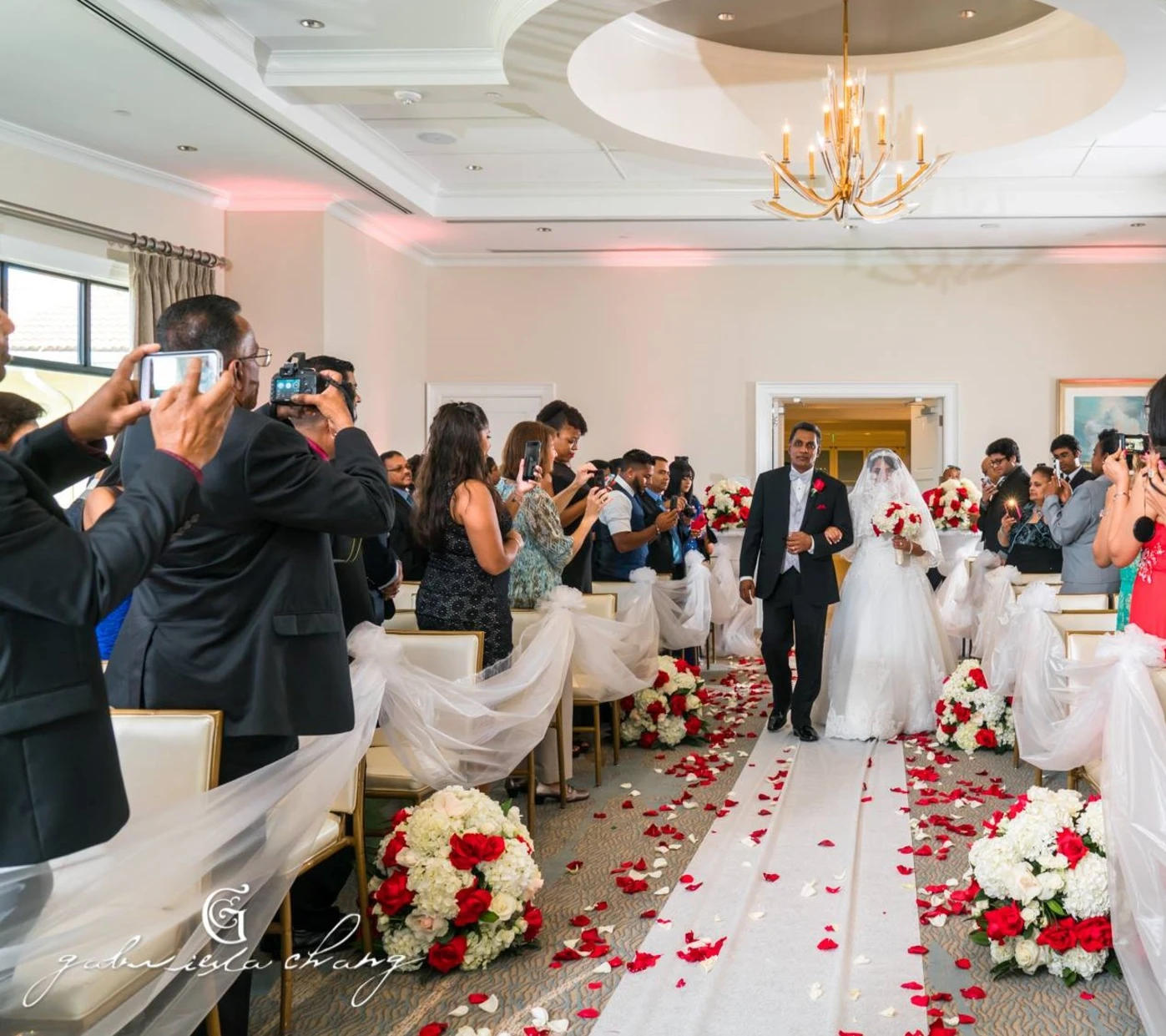 Bride in a white wedding gown and veil walking down a petal-strewn aisle on the arm of a man in a tuxedo, with guests on both sides taking photos and watching in a decorated indoor venue.