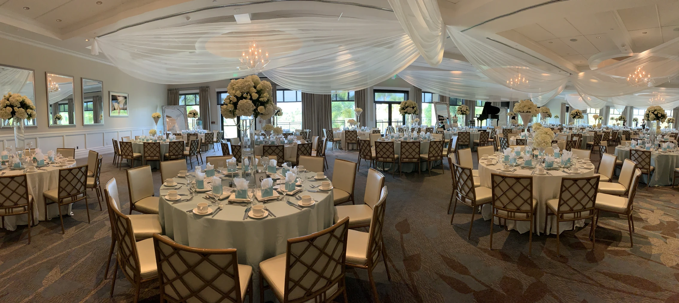 Elegant banquet hall set up with round tables covered in light blue tablecloths, each adorned with white floral centerpieces and formal place settings, under soft white draped ceiling fabric and chandeliers.