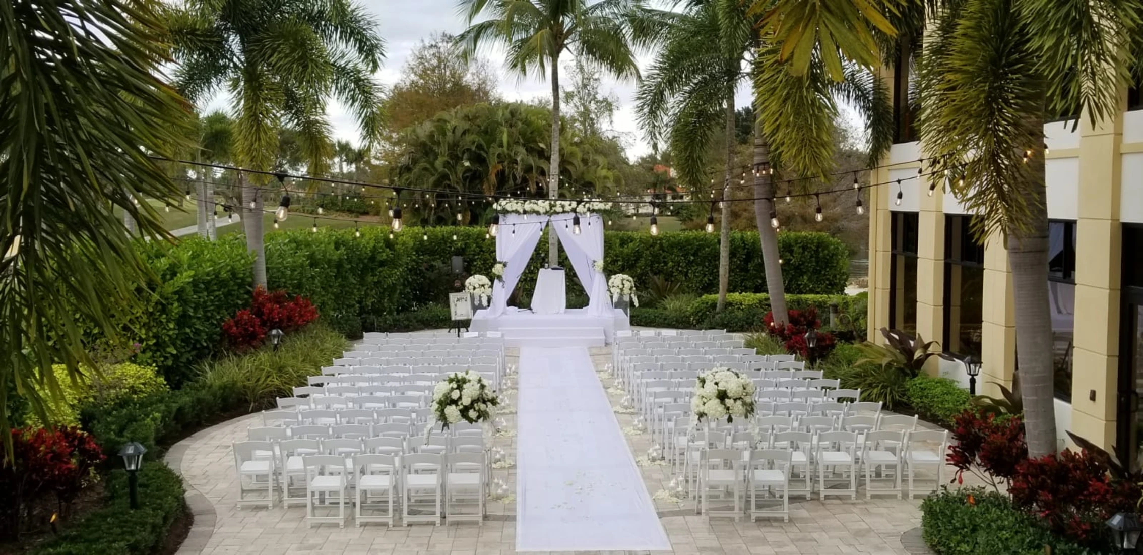 Outdoor wedding ceremony setup with rows of white chairs flanking a white aisle runner leading to a white floral arch decorated with flowers, surrounded by palm trees and garden greenery under string lights.