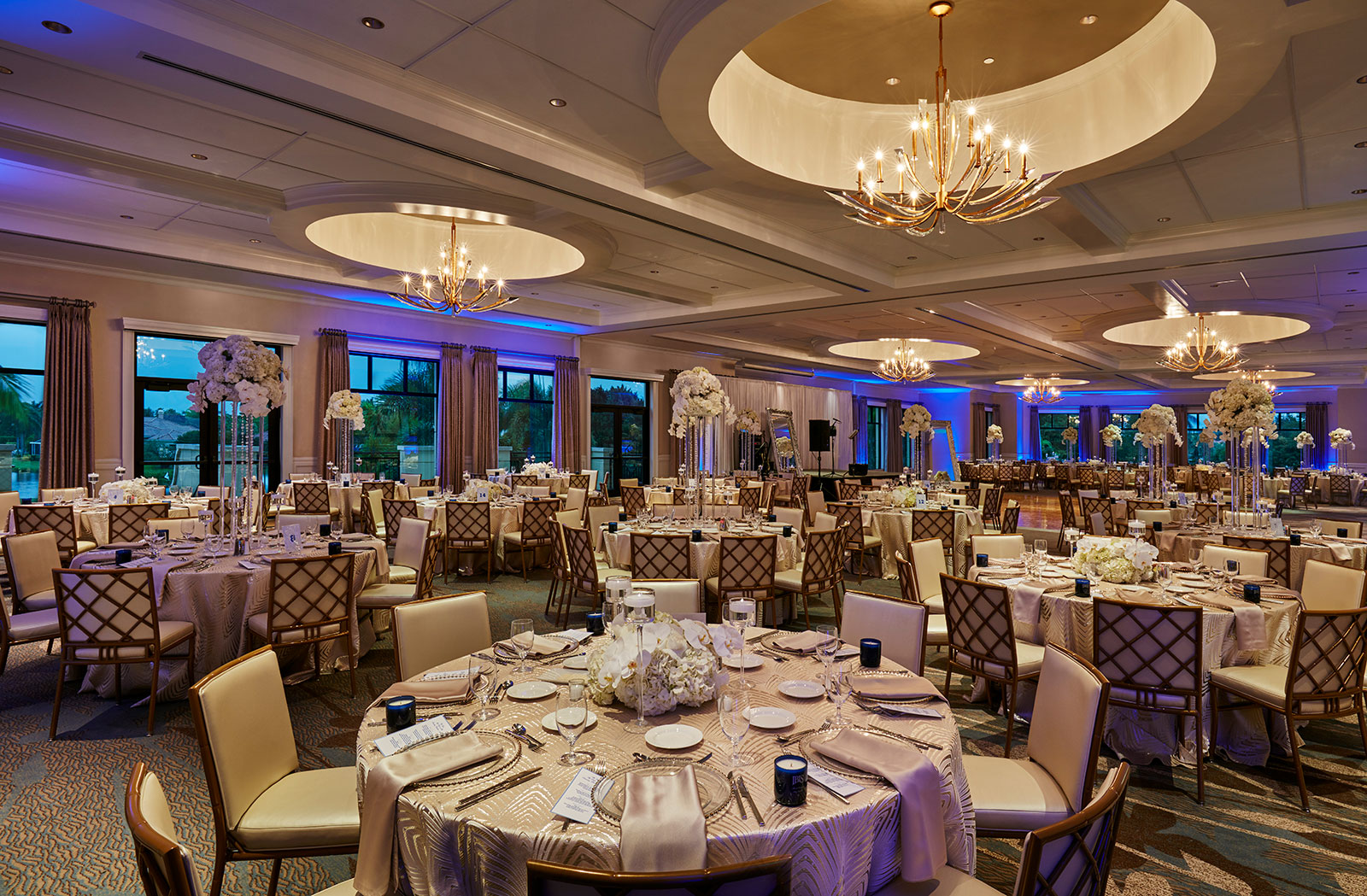 A large elegant ballroom set for a gala with round tables covered in gold-patterned tablecloths, cream chairs, floral centerpieces of white flowers on tall stands, neatly arranged glassware, silverware, and napkins under soft warm lighting with chandeliers and blue accent lights along the walls.