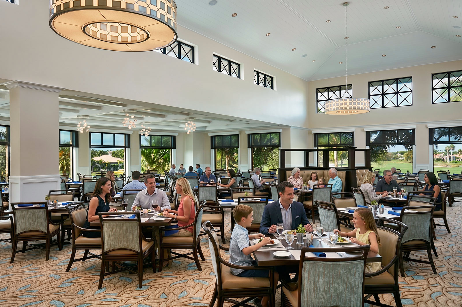 Spacious, elegant dining room with large windows showing greenery outside, filled with people seated at tables eating and conversing, featuring modern light fixtures, beige walls, and patterned carpet.