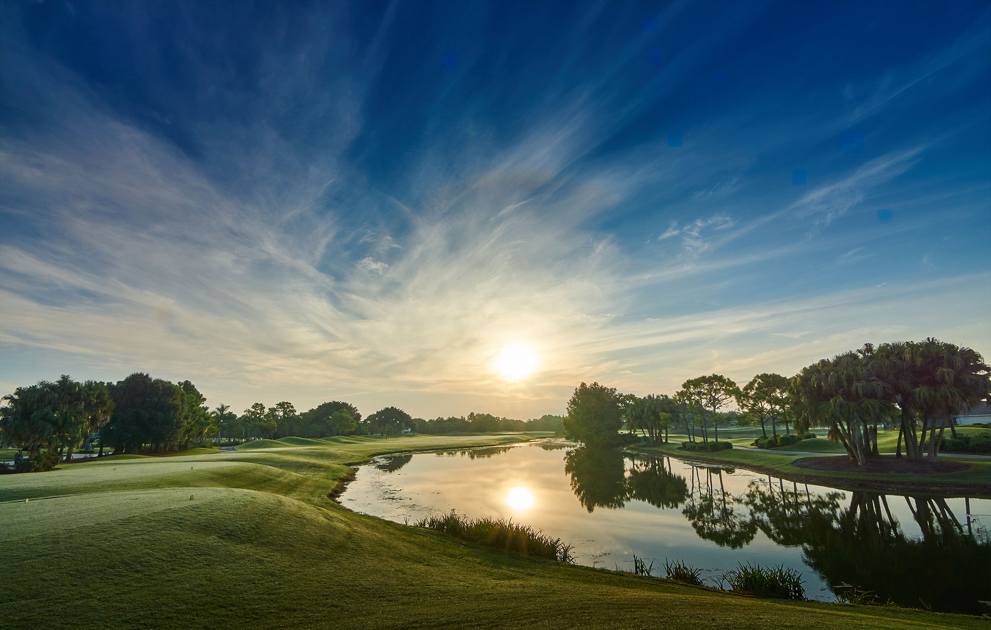 Sunrise over a golf course with a calm pond reflecting trees and a sky filled with wispy clouds.