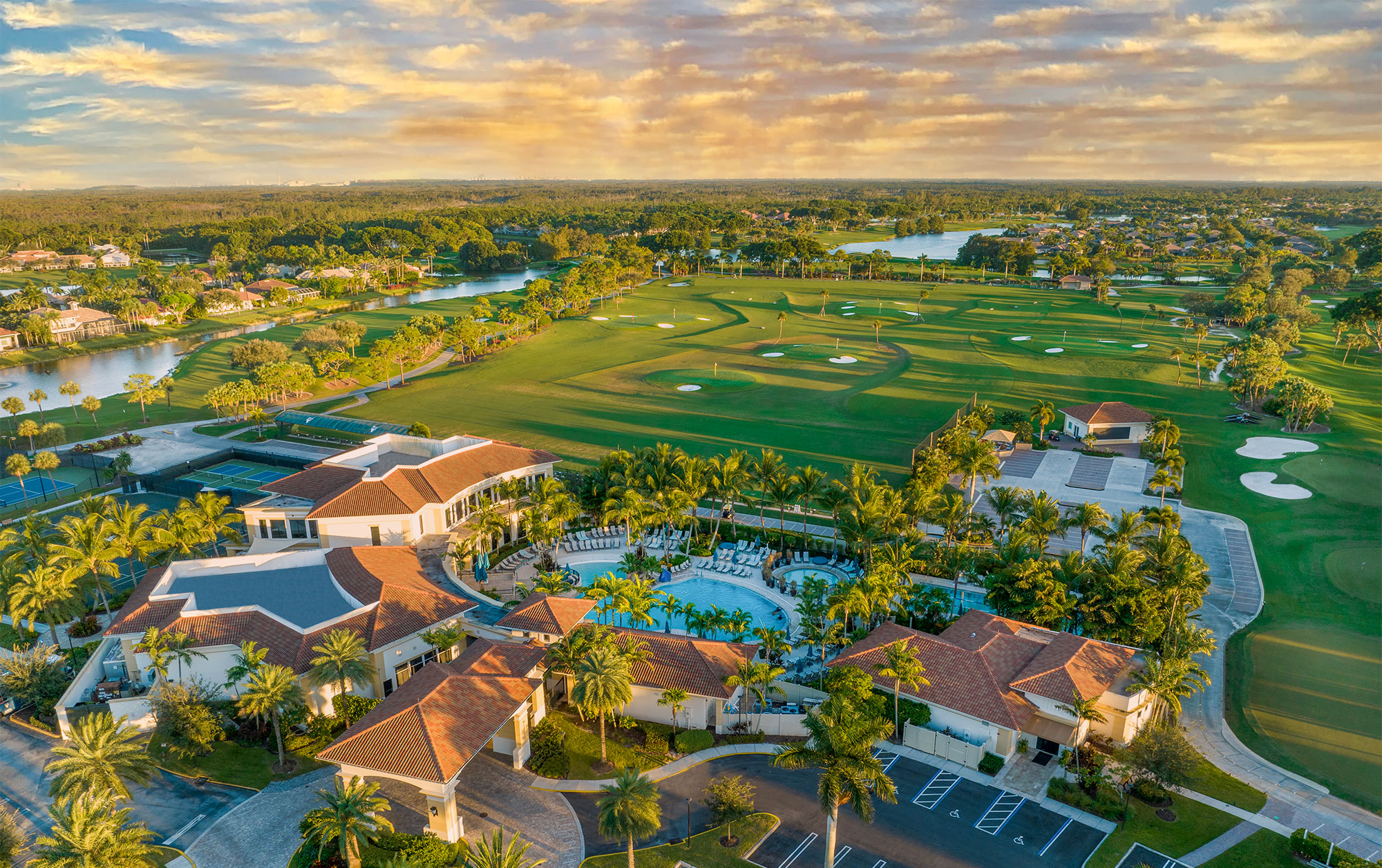 Aerial view of a luxury resort with red-tiled roofs, a large pool surrounded by palm trees, adjacent to a golf course with sand bunkers, green fairways, and small ponds under a partly cloudy sky at sunset.