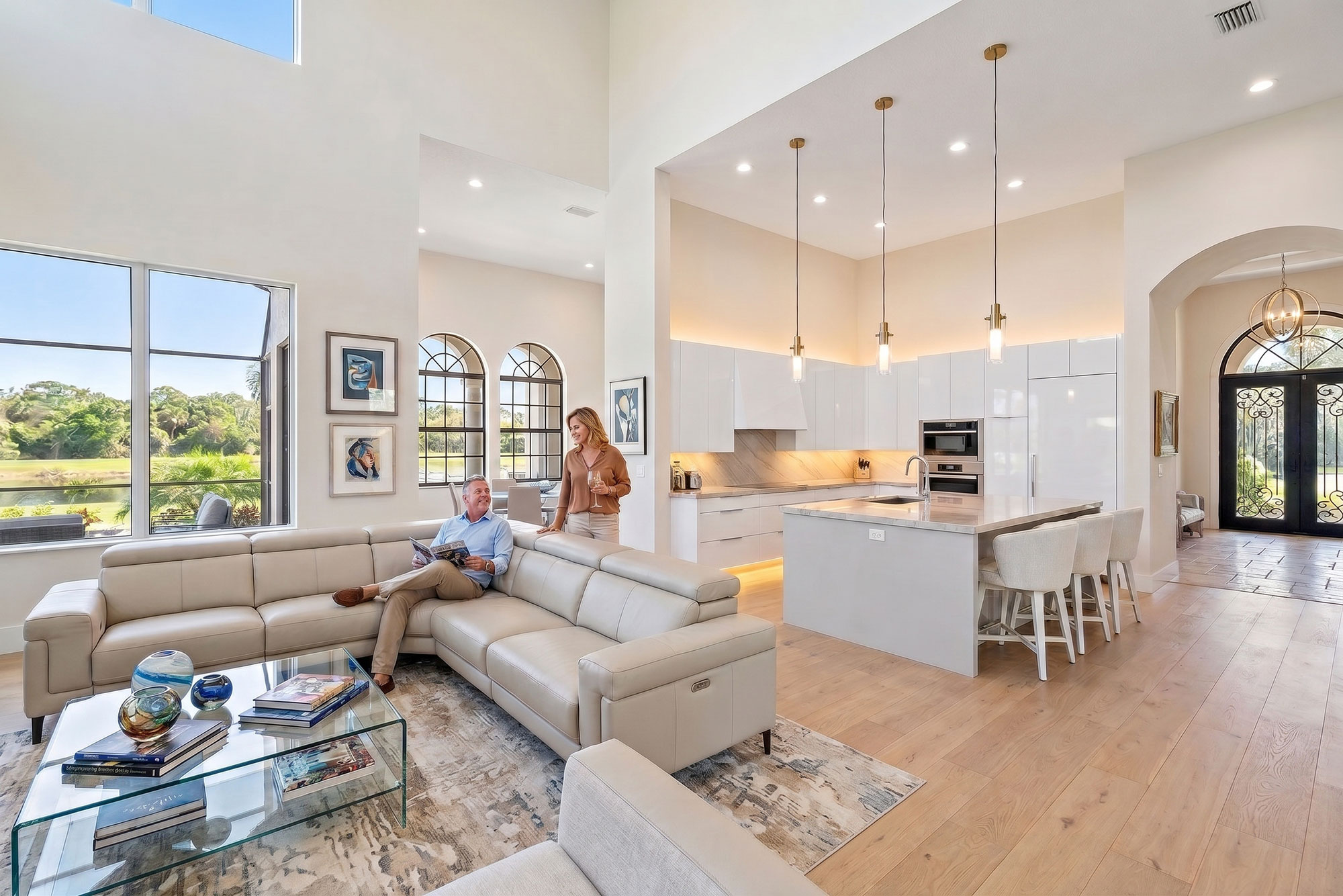 Bright, modern open living space with beige leather sectional sofa, glass coffee table with decorative items, and a kitchen with white cabinetry, island, and pendant lights; a man sitting on the sofa reading a magazine looks up at a woman standing beside him holding a wine glass; large windows show green outdoor scenery.