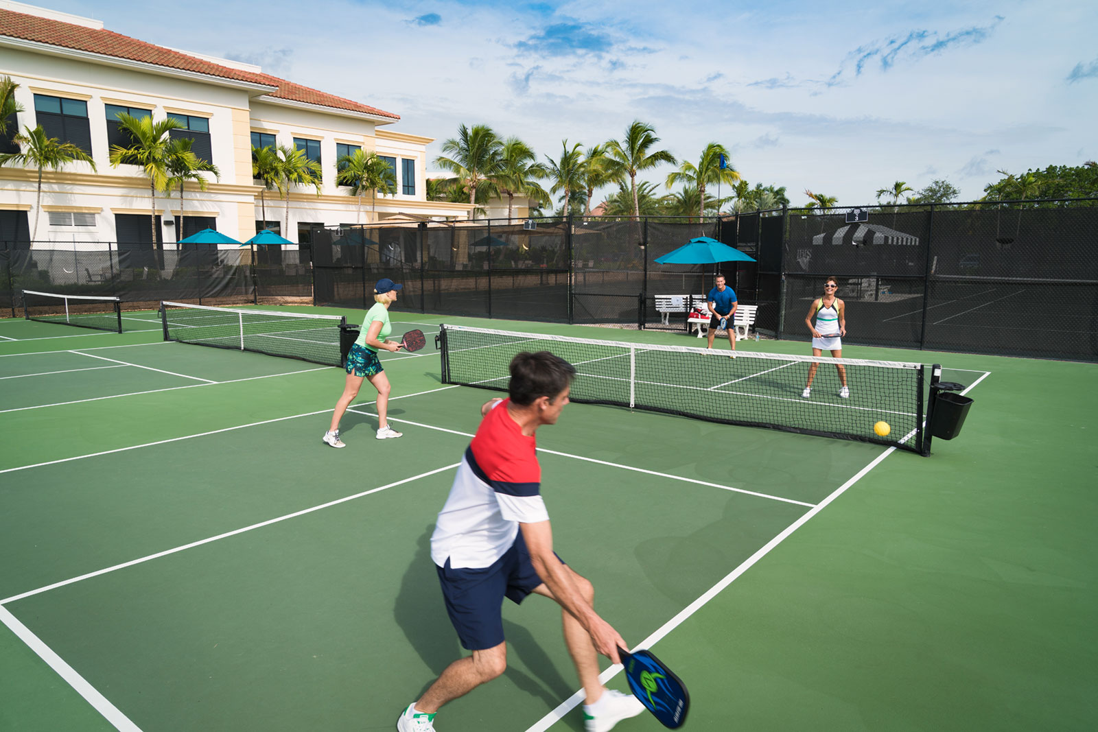 Four adults playing doubles pickleball on an outdoor court with a green surface, surrounded by palm trees and a building in the background, the yellow ball is near the net.