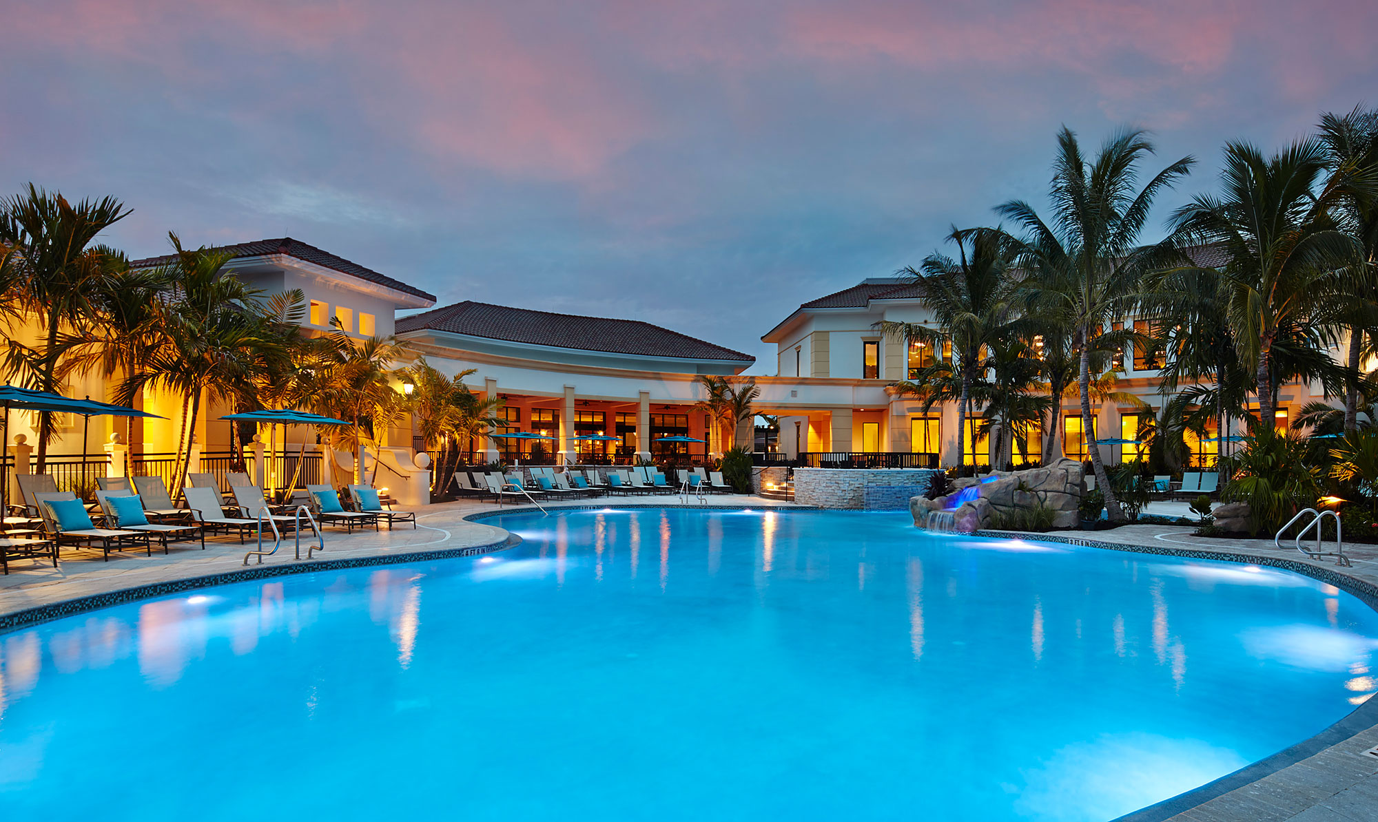 Large, illuminated outdoor swimming pool at twilight with lounge chairs and blue umbrellas along the deck, surrounded by palm trees and a yellow-lit resort building in the background.