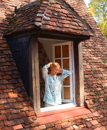Jeune femme blonde en pyjama souriante, prenant l'air à travers une fenêtre de toit en tuiles. Les Hauts de Loire – Hôtel, Restaurant & Spa – 5 étoiles.